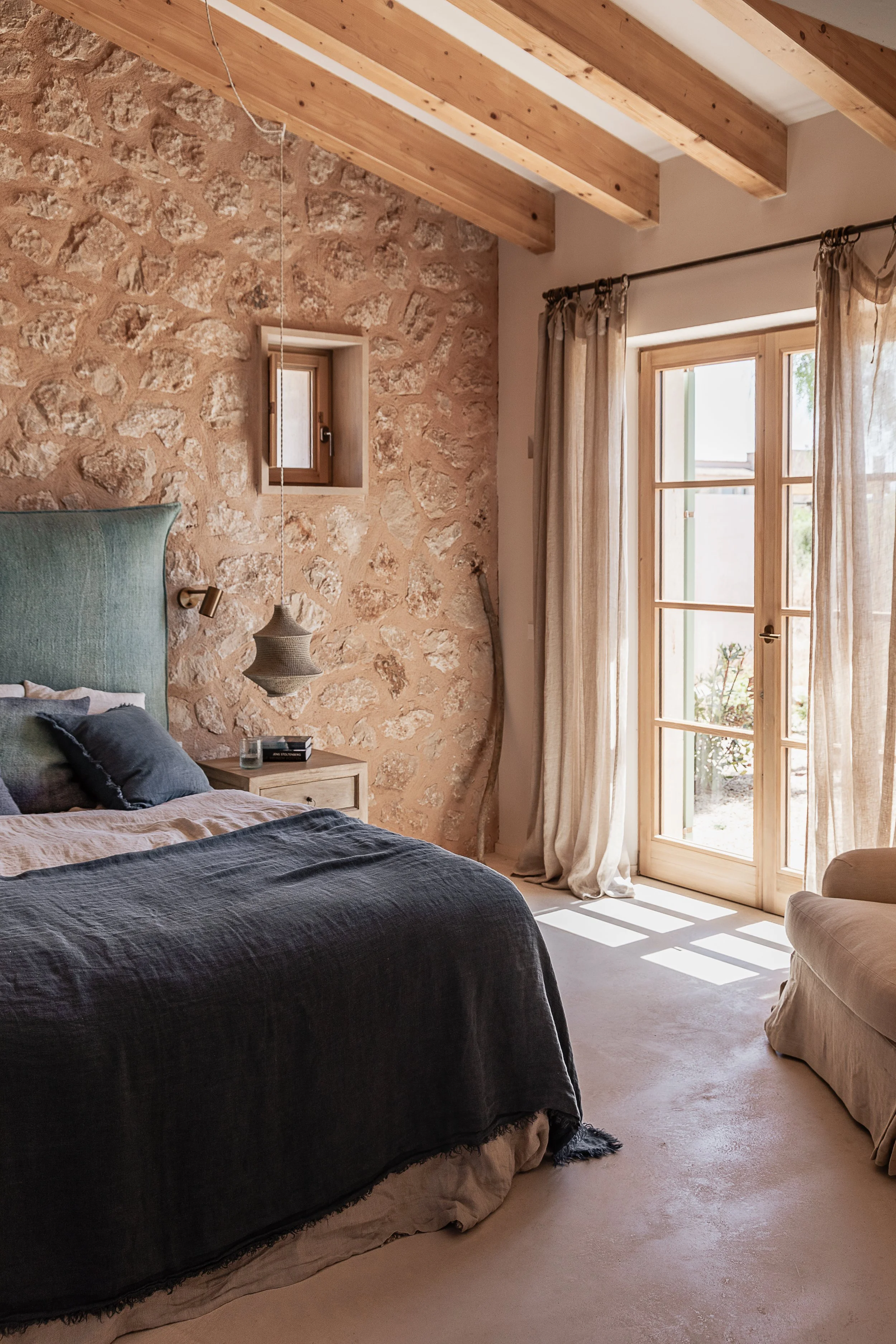 Bedroom with exposed timber beams, natural stone wall, linen curtains and French doors opening onto the courtyard of a Mediterranean home in Mallorca.