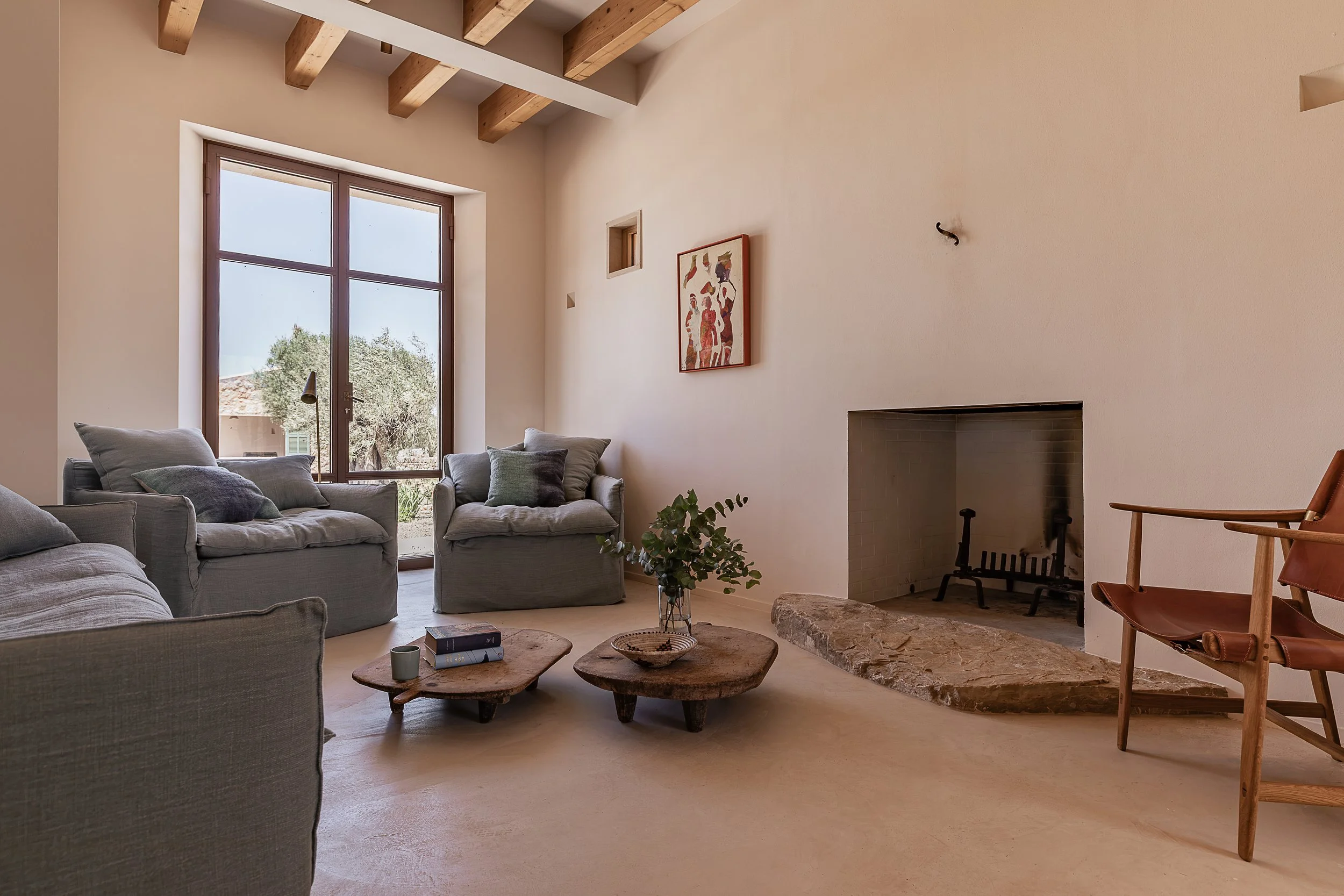 Living room with a fireplace featuring a hand-selected Binissalem stone hearth, exposed timber beams and seamless microcement flooring in a Mediterranean home in Mallorca.