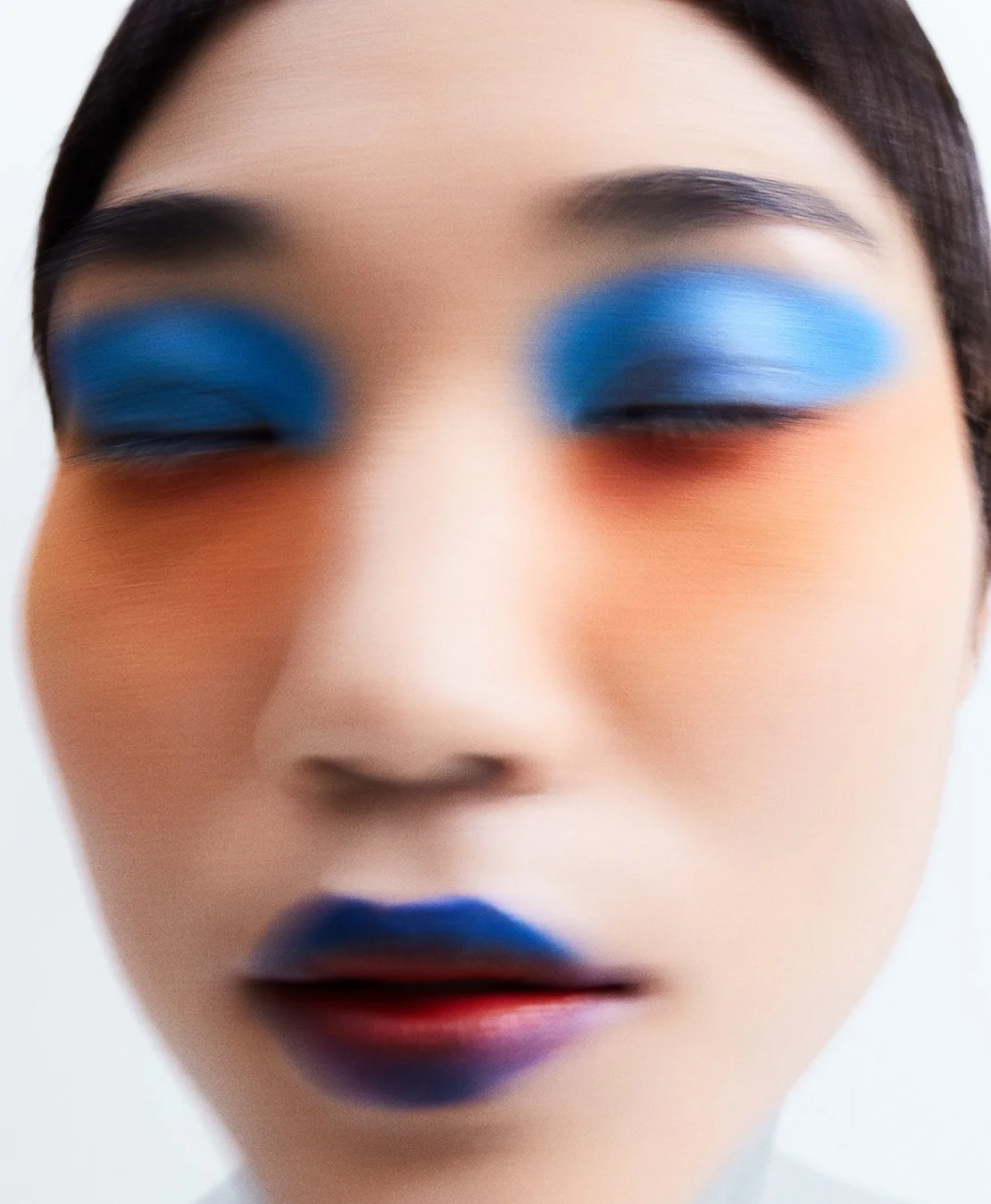 Close-up portrait of a woman with colorful makeup, including bold blue eyeshadow and lipstick, blurred to create an artistic effect.