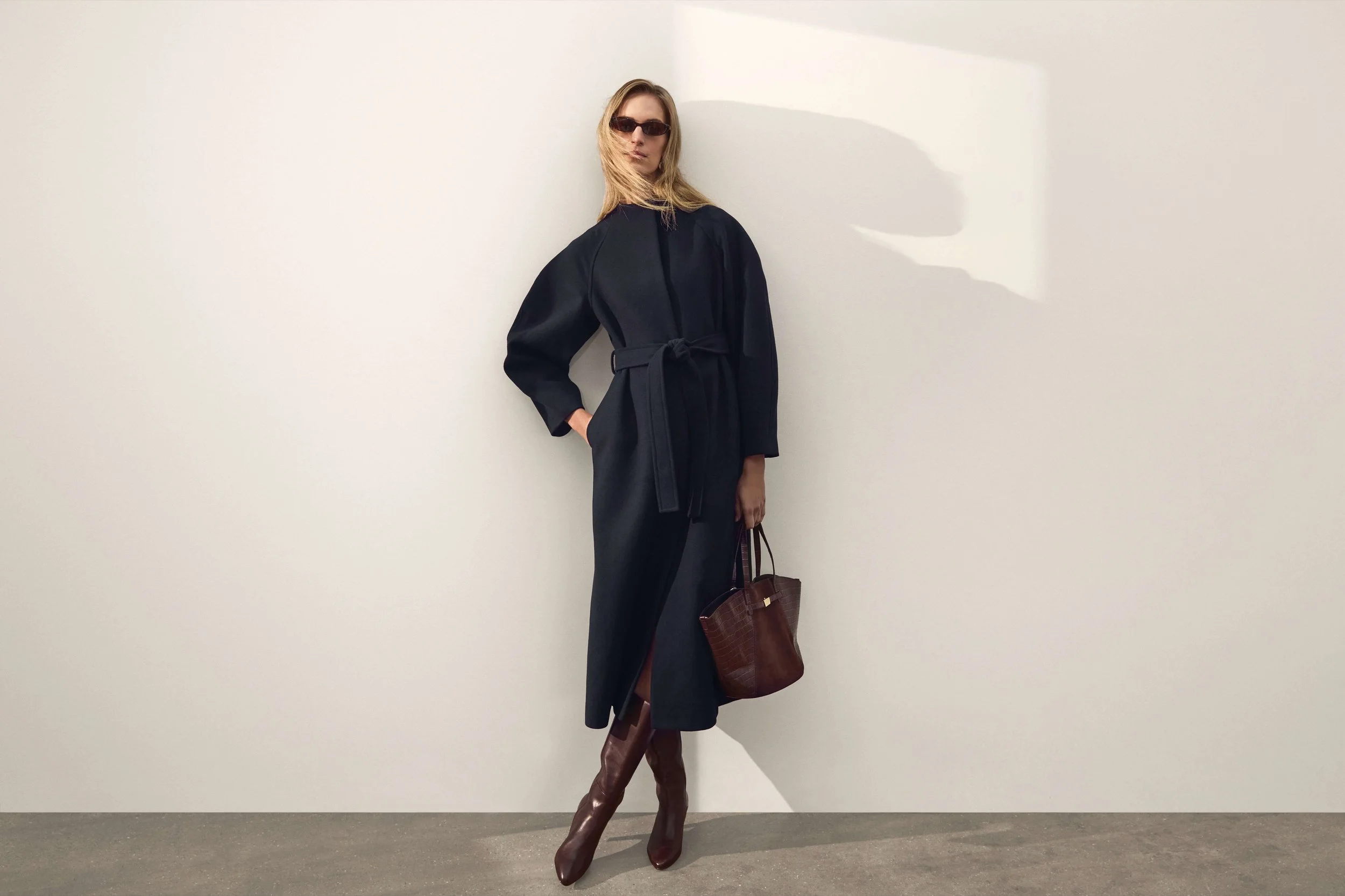 A woman with blonde hair wearing black sunglasses, a black belted long-sleeve dress, brown knee-high boots, and carrying a brown handbag, standing against a white wall with her left hand in her pocket and her right hand holding the handbag.