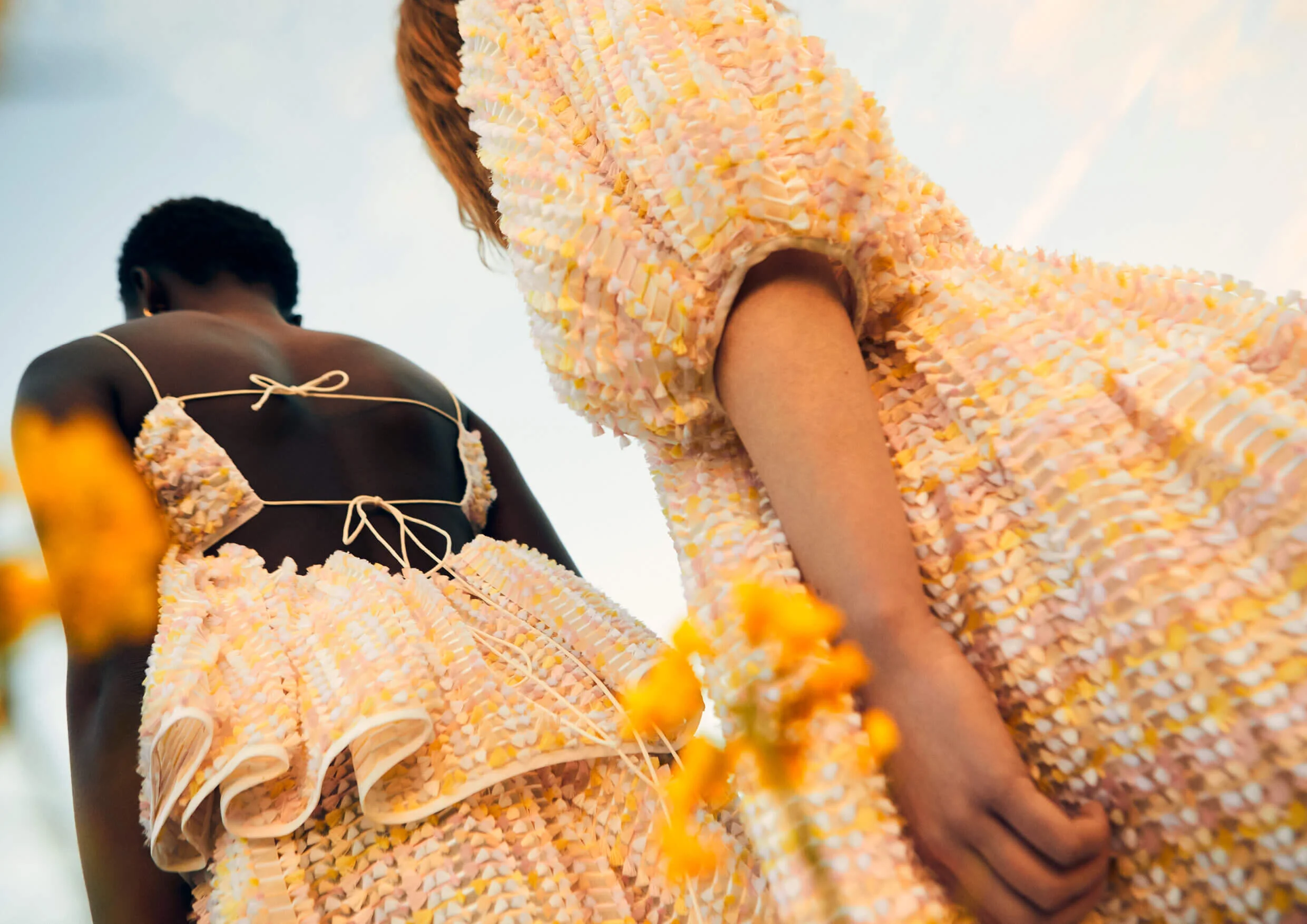Close-up of two women wearing colorful, textured dresses, viewed from below against a sky background.