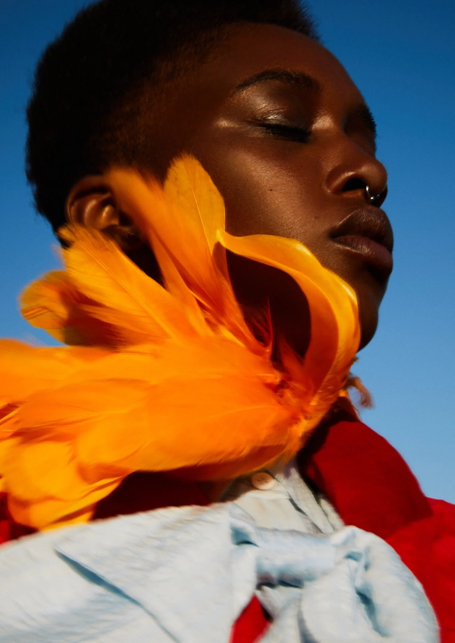 A person with dark skin and short hair, wearing a sequin face mask and a yellow feathered garment, with a bright blue background.