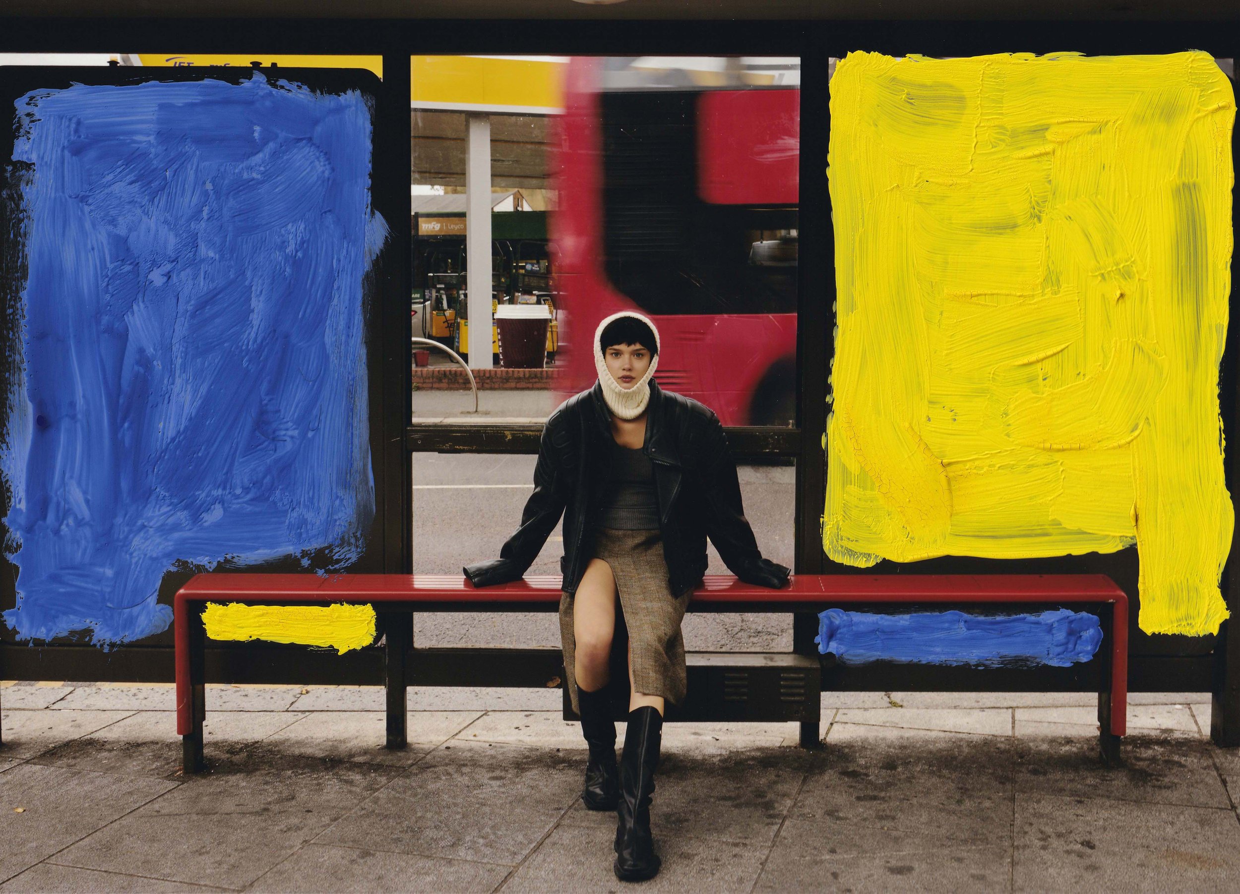 A woman with short dark hair, wearing a white knit hat, black leather jacket, and a beige skirt, sitting on a red bench at a bus stop with painted blue and yellow squares on the glass panels around her.