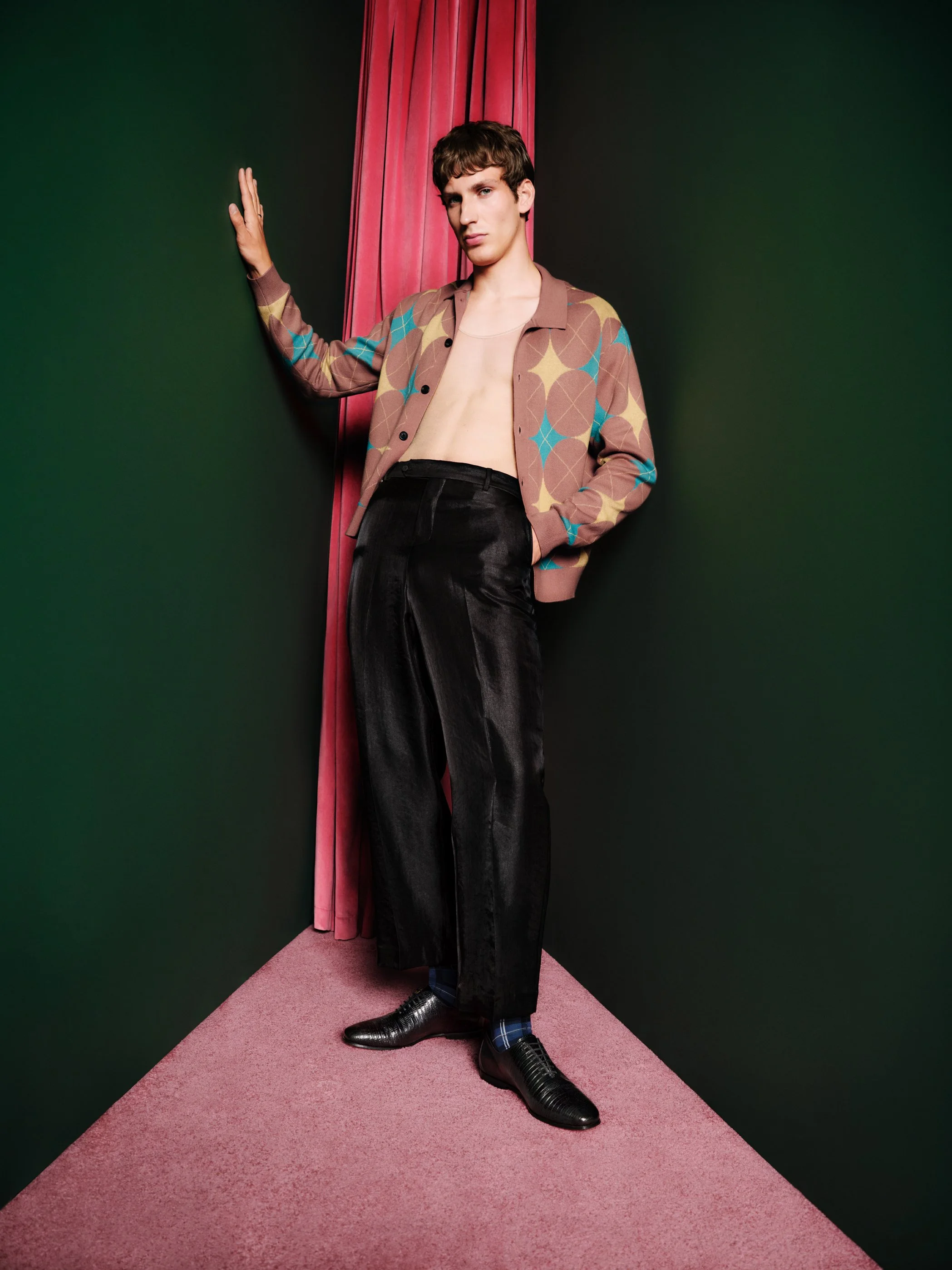 A young man with a leather jacket, black trousers, and shiny shoes standing on a pink carpet with green and dark gray walls, leaning against the wall with one hand.
