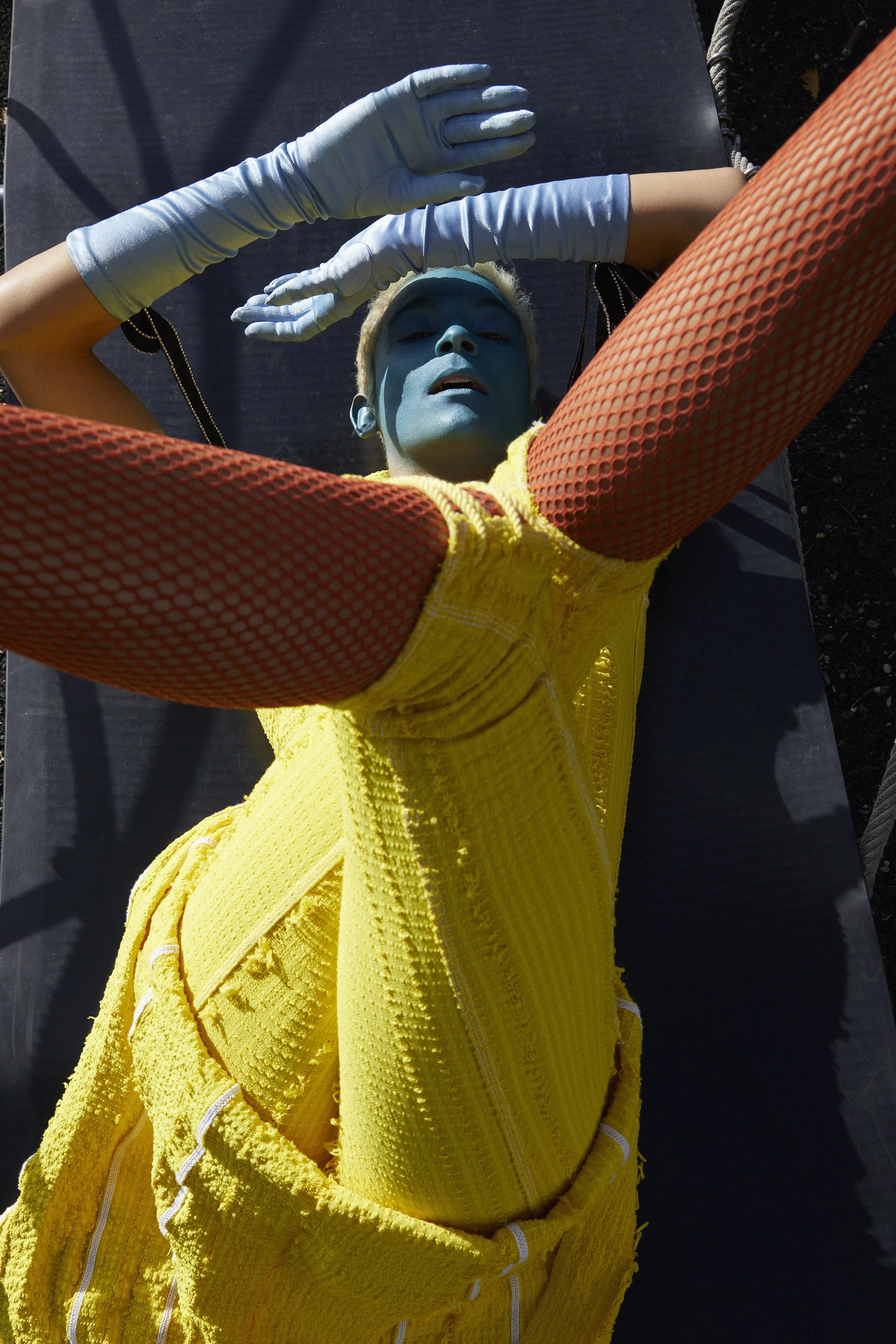 Person with blue face paint, wearing bright yellow textured clothing with white accents, lying on a black surface with arms raised and wearing long white gloves.