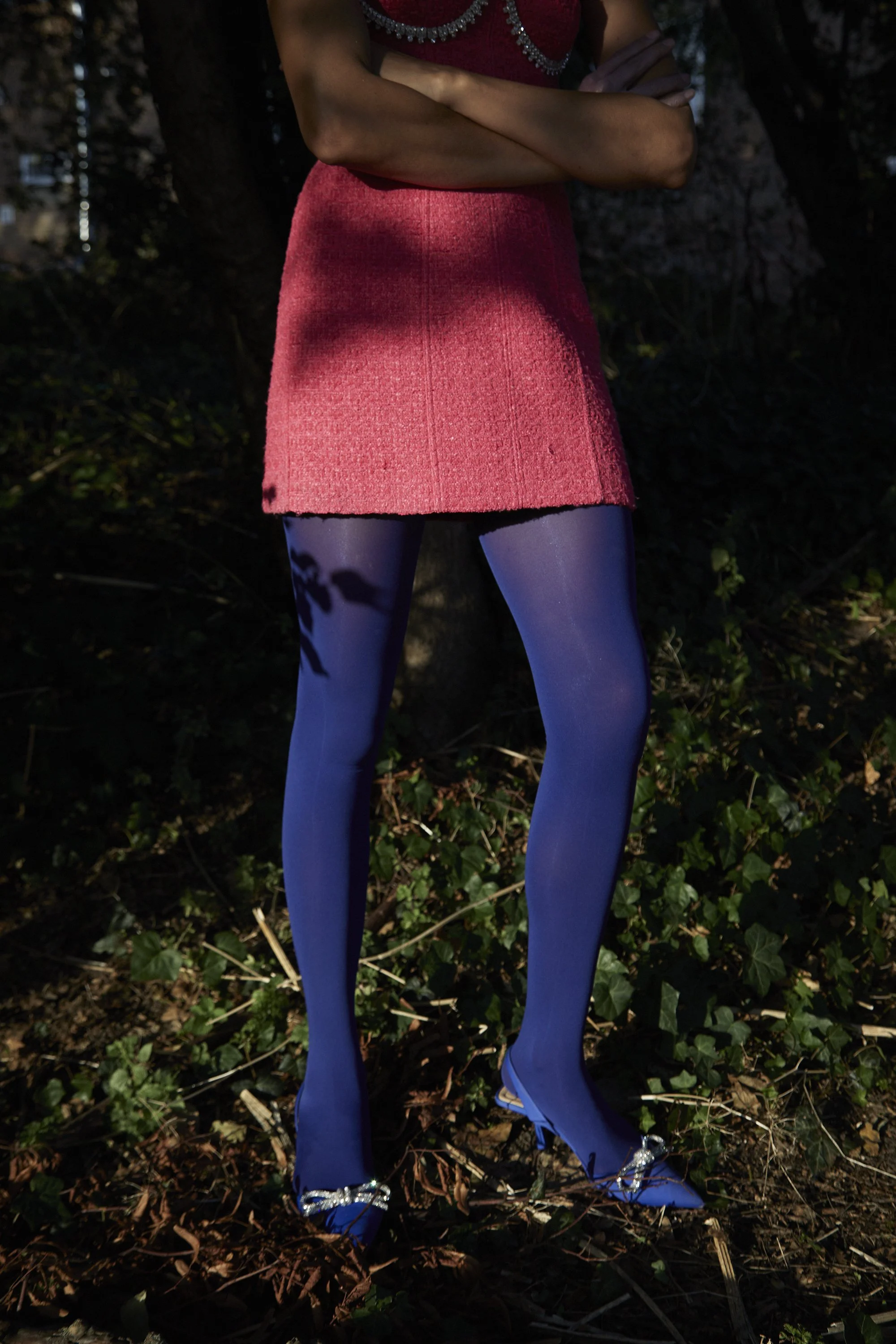 A pair of blue mannequin legs with sparkly heels, wearing a red dress with silver embellishments, standing outdoors on dirt and fallen leaves at night.