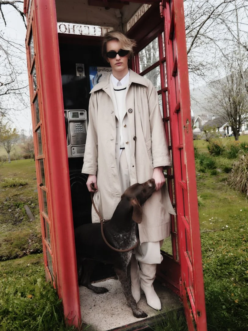 A woman in sunglasses and a beige trench coat standing inside a red British-style phone booth. A large brown and black dog is standing beside her, licking her hand. The phone booth contains a telephone and a poster inside, with a park and trees visible outside.