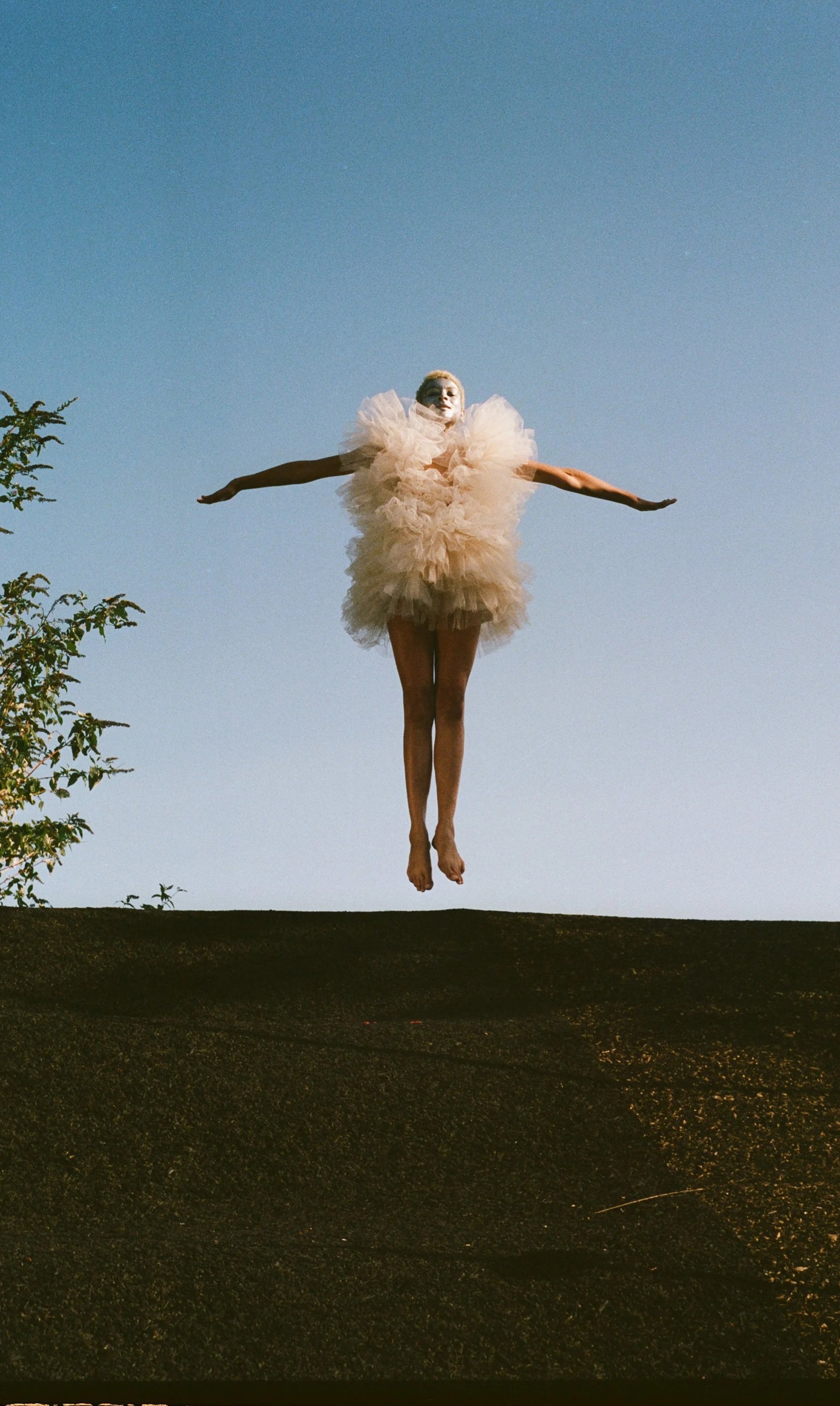 A person dressed in a fluffy, beige tutu-like outfit is floating or jumping in the air against a clear blue sky with a few trees visible on the left side.