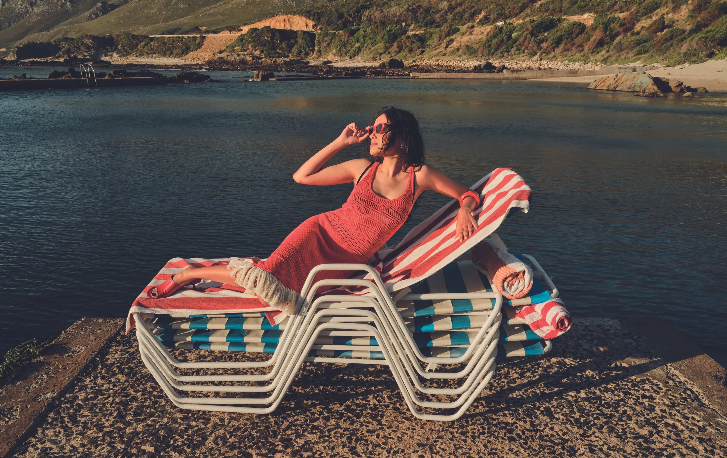 A woman relaxing on a lounge chair by the water at a beach, wearing sunglasses and a red dress, with stacked lounge chairs and a rocky shoreline in the background.