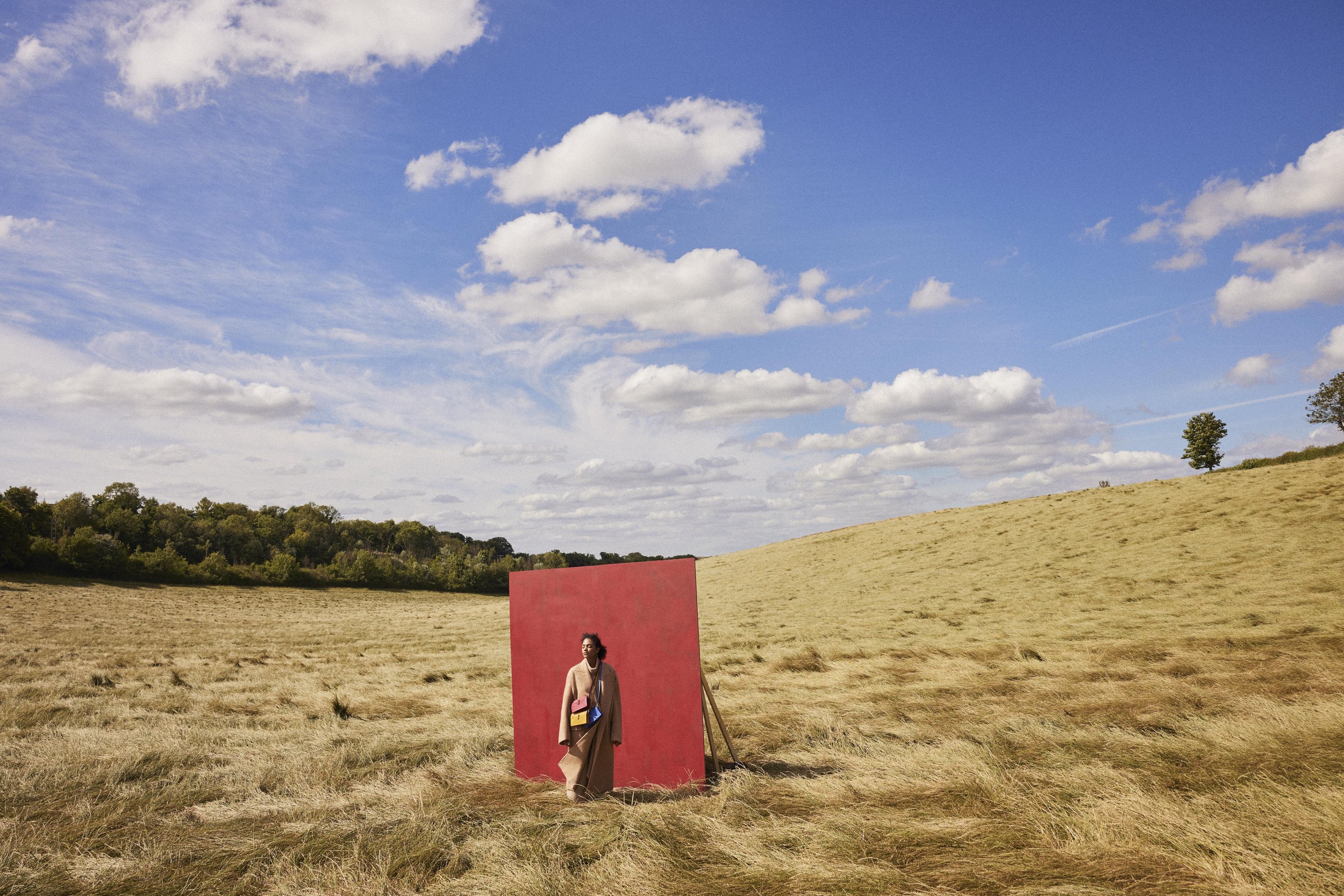 An outdoor scene with a vast open field, a few trees, and a blue sky with scattered clouds. There is a large red panel in the field standing as a backdrop behind a woman with her hair tied back, wearing a beige coat.