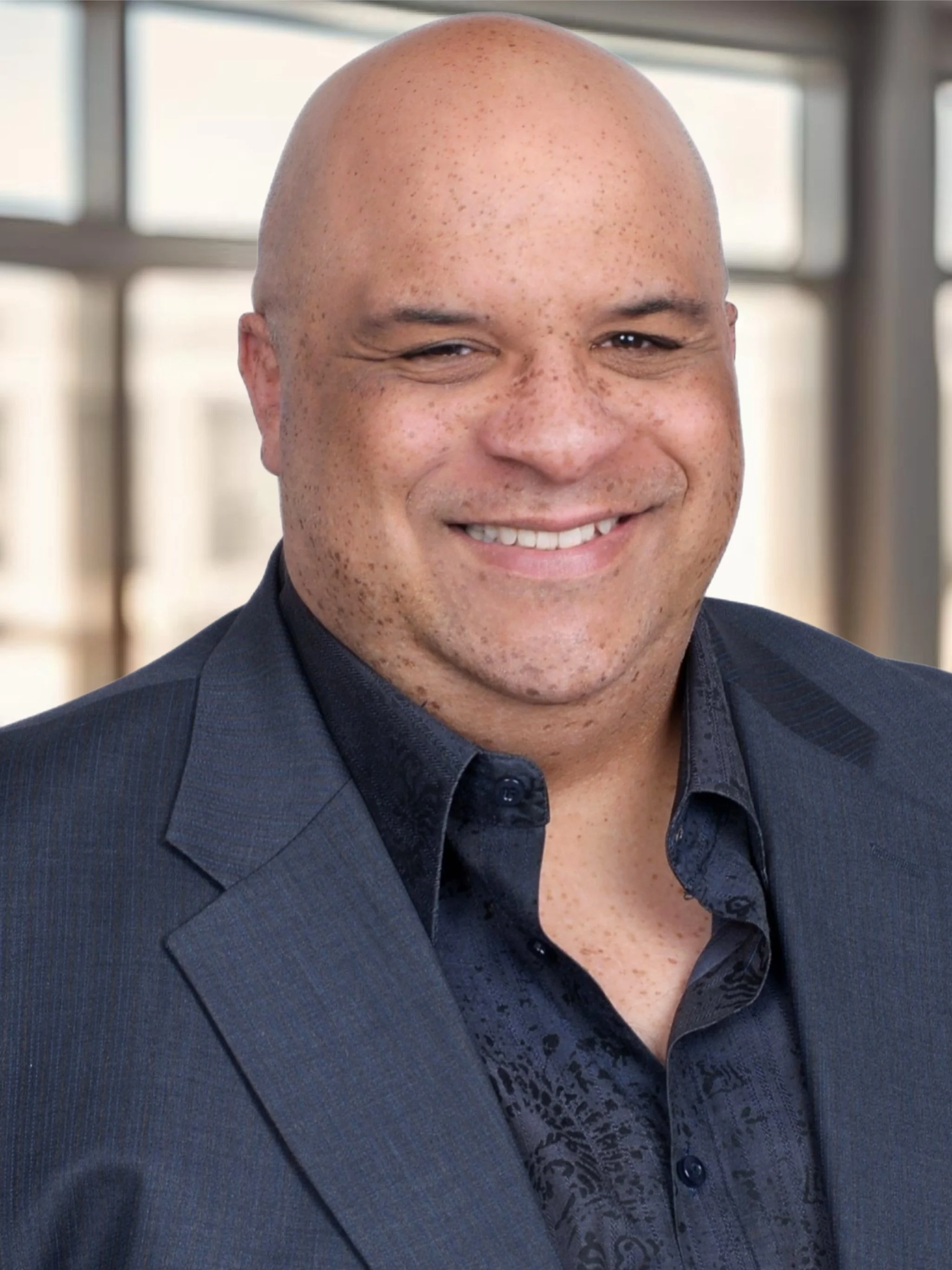 A man with a bald head and freckles, smiling, wearing a dark suit and patterned shirt in an indoor setting with large windows in the background.
