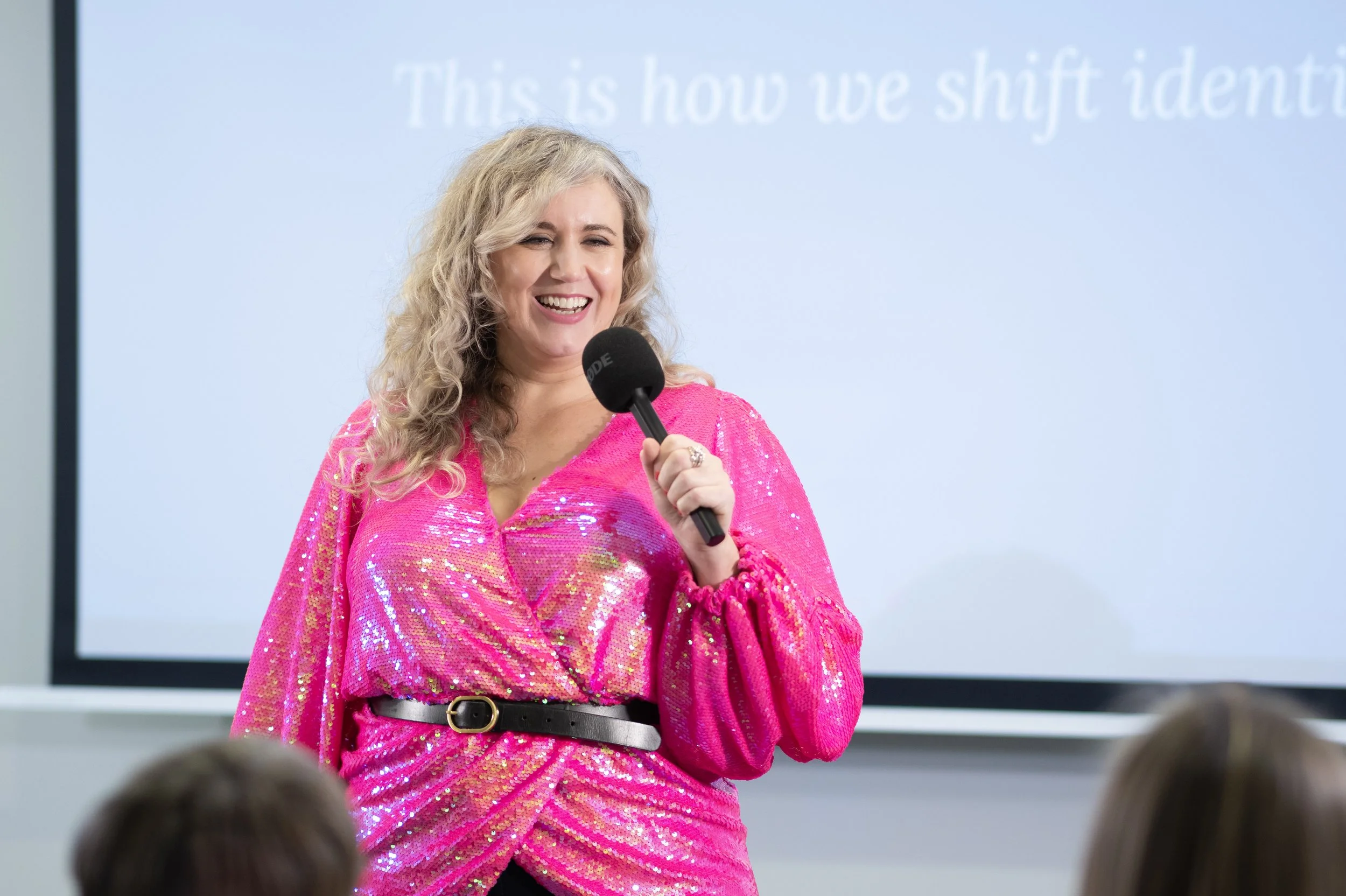 A woman with blonde curly hair wearing a bright pink, shiny, long-sleeved top and black belt, smiling and holding a microphone in front of an audience.