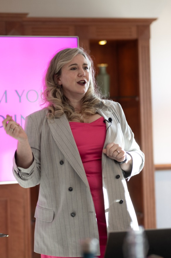 A woman with blonde hair wearing a pink dress and a gray pinstripe blazer is speaking in front of a pink screen, gesturing with her left hand during a presentation.
