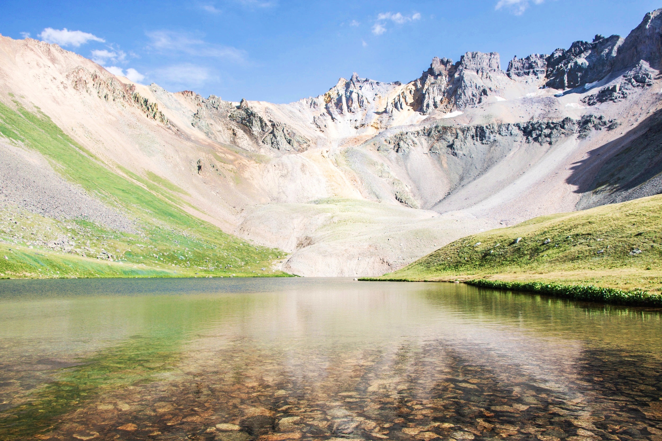 Scenic mountain landscape with a clear lake in the foreground, rocky peaks and green slopes under a blue sky.