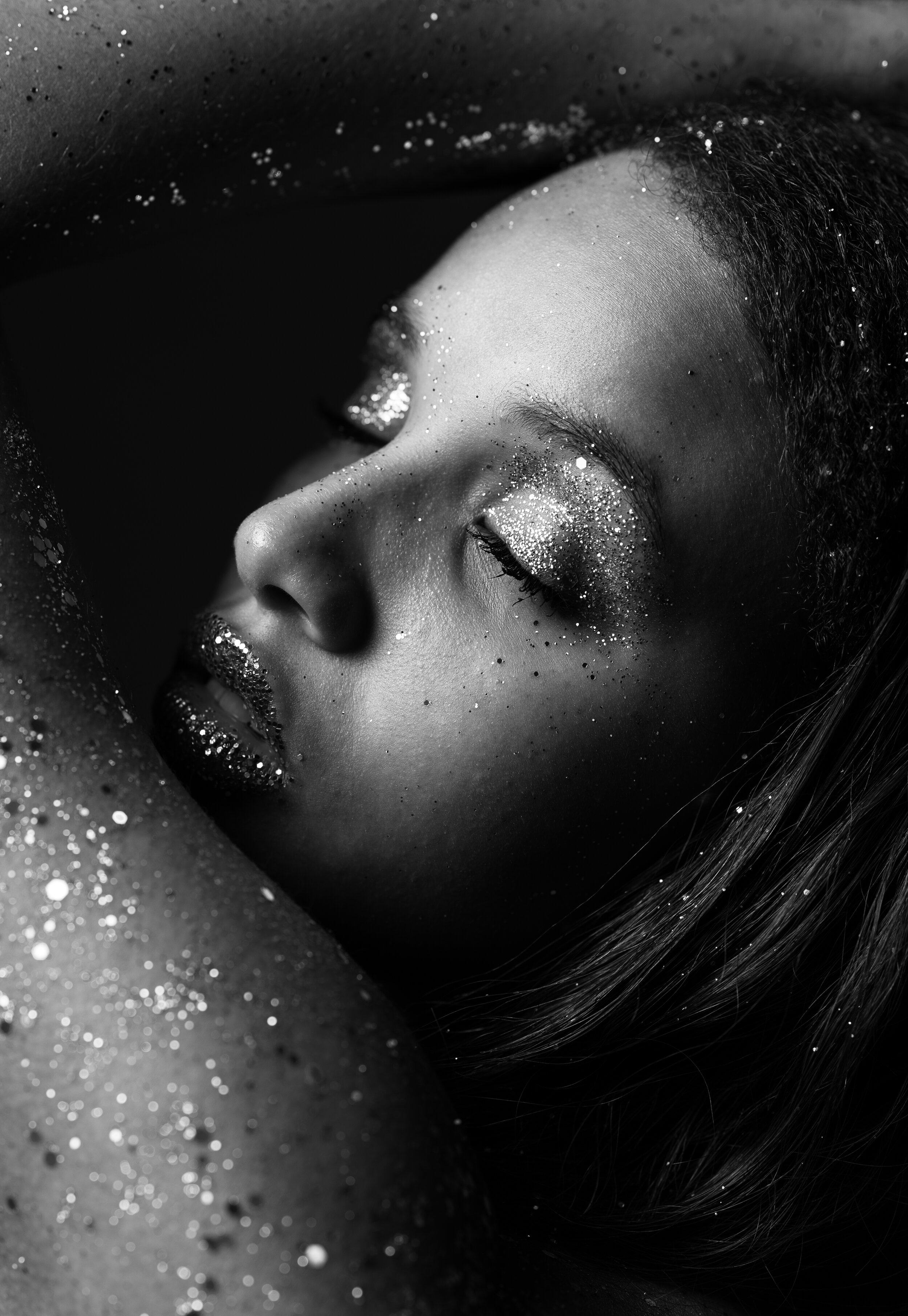 Black and white close-up of a person with closed eyes, covered in glitter, with an artistic pose.
