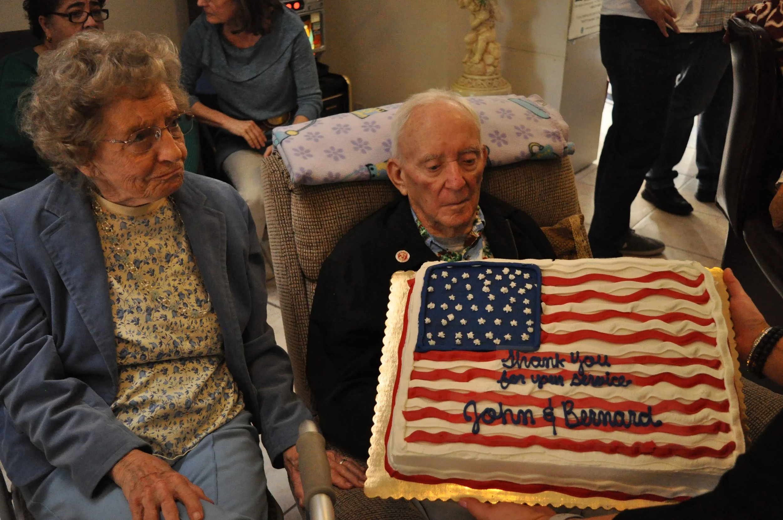 Two elderly women and an elderly man sitting at a table with a cake decorated with an American flag pattern, with a message of thanks for their service, likely at a celebration or event. In the background, there are other people standing and sitting.