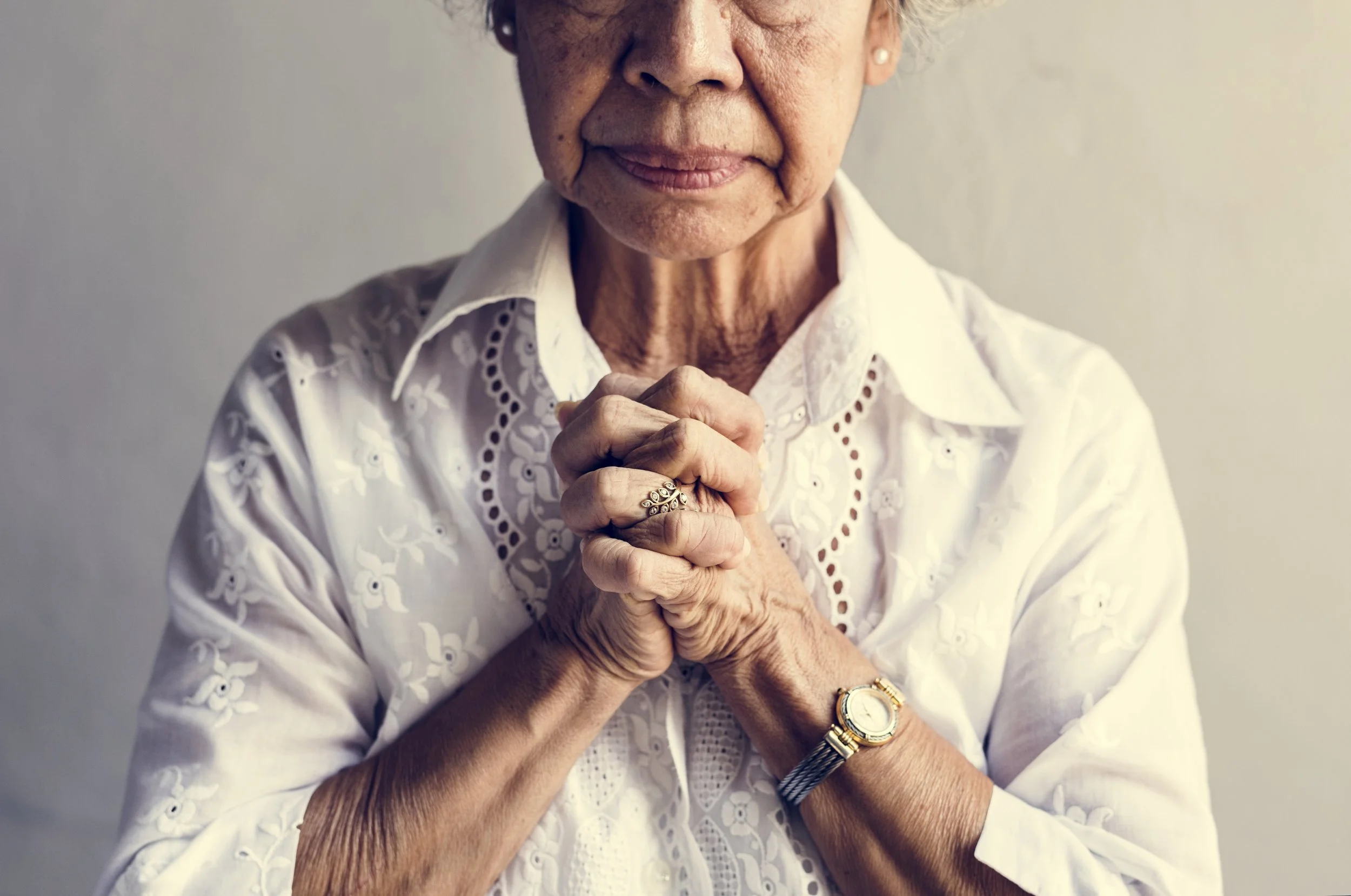 Close-up of an elderly woman with hands clasped together, wearing a white embroidered blouse and watch, with a neutral background.