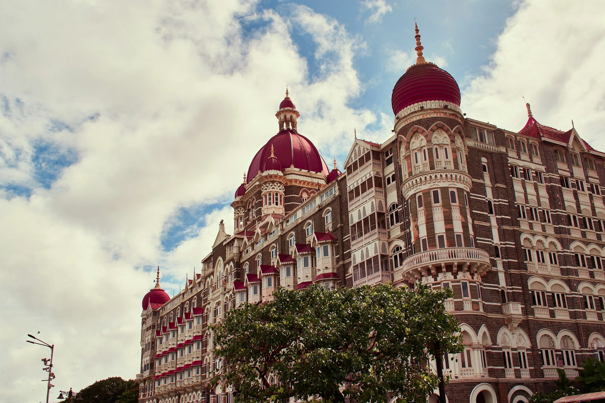A large historic building with multiple red domes and intricate architectural details, set against a partly cloudy sky, with some trees and streetlights in the foreground.