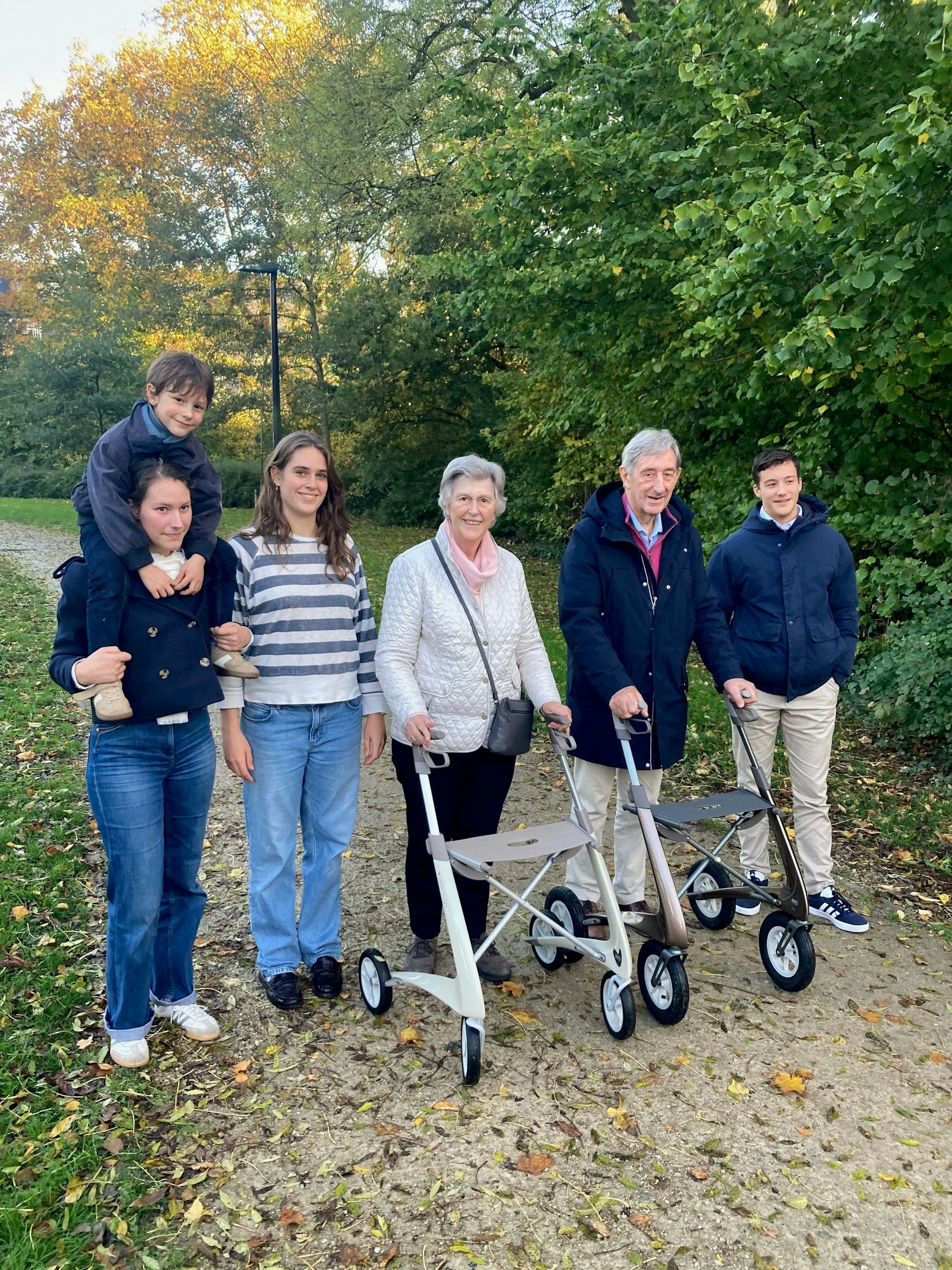 A multigenerational family of six people, including children and elderly, walking together outdoors on a fall day surrounded by green and yellow trees, with some of the group using walkers.