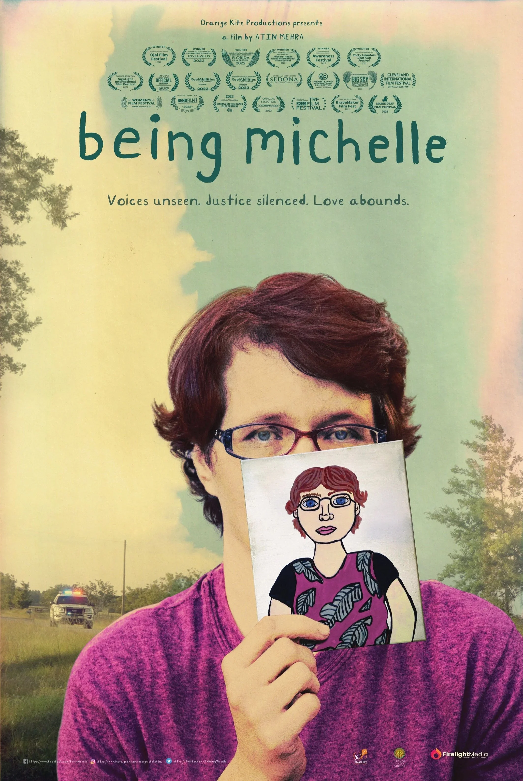 A woman with short reddish hair and glasses holding a cartoon drawing of herself in front of her face. The background features a grassy field, a police vehicle, trees, and a colorful sky with a gradient from yellow to pink.