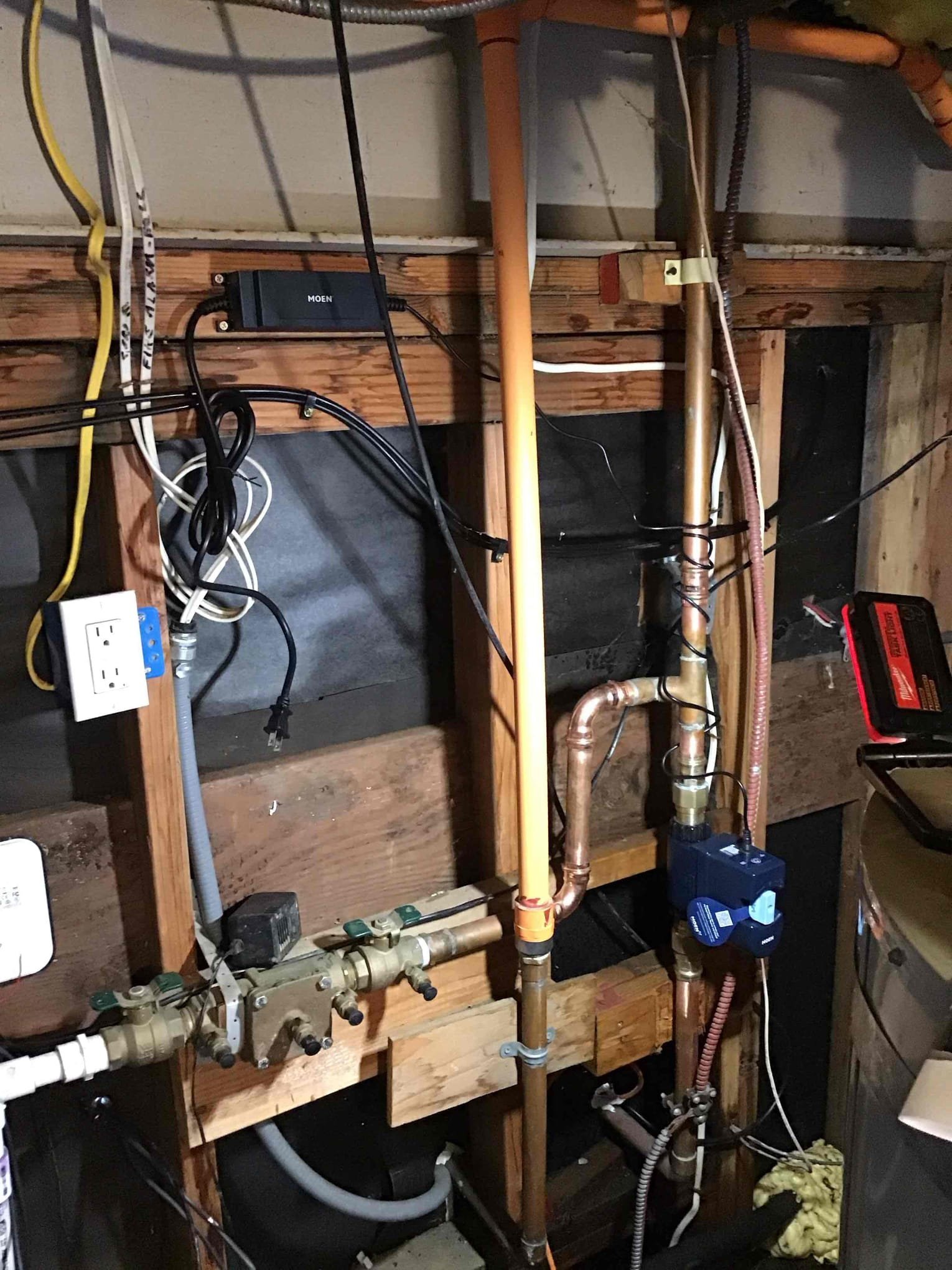 The image shows an unfinished wall with exposed copper and PVC pipes, electrical wiring, and some electronic components, indicating a plumbing and electrical setup in a construction or renovation area.