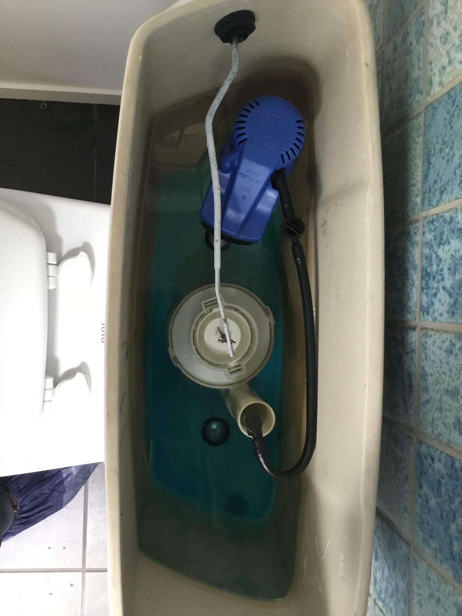 A beige toilet tank with water inside, containing a blue float valve and a black and gray fill valve, with a white and blue tiled floor in the background.