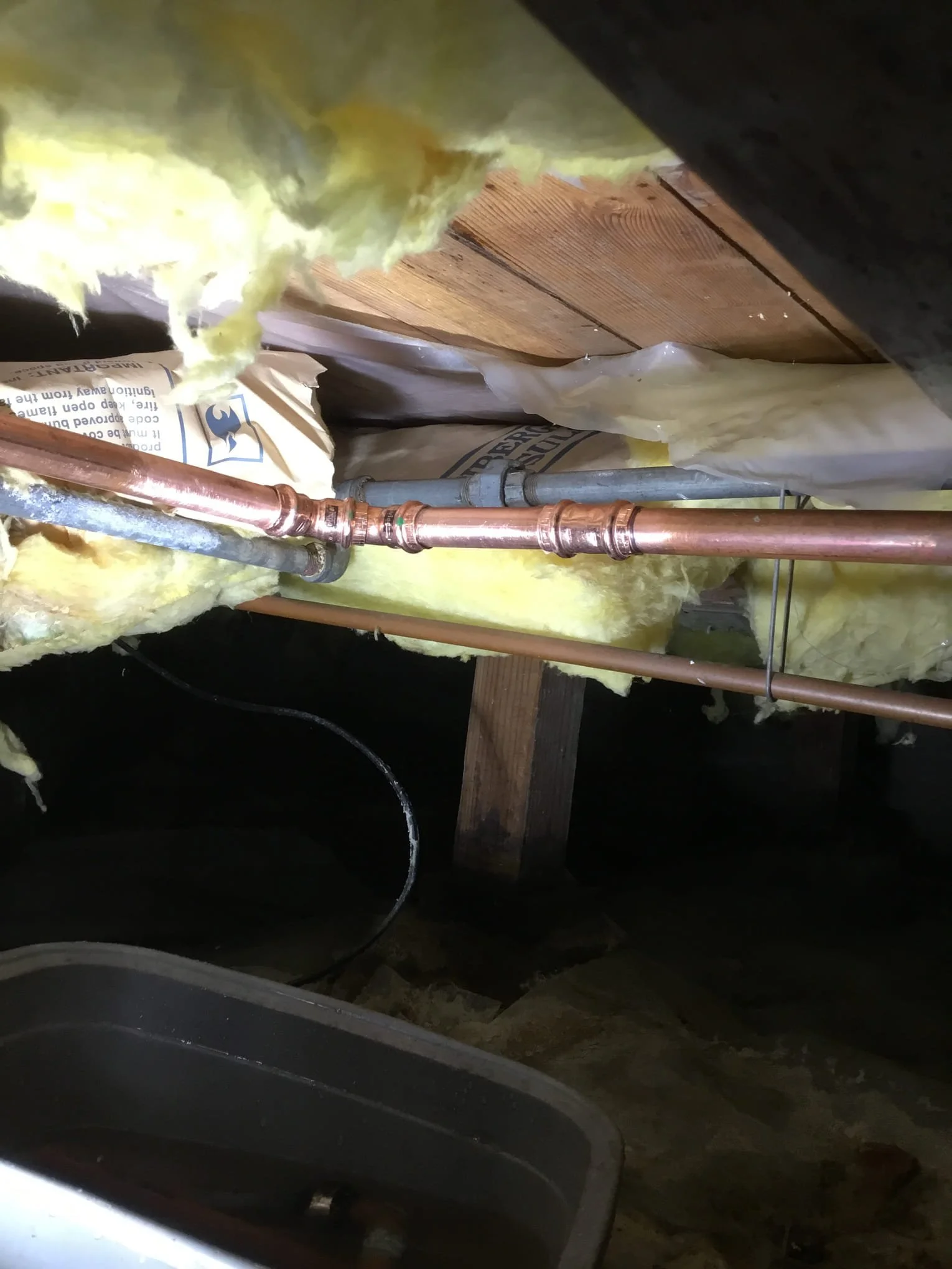 Under an attic or crawl space, showing insulation material, copper piping, and some wooden beams.