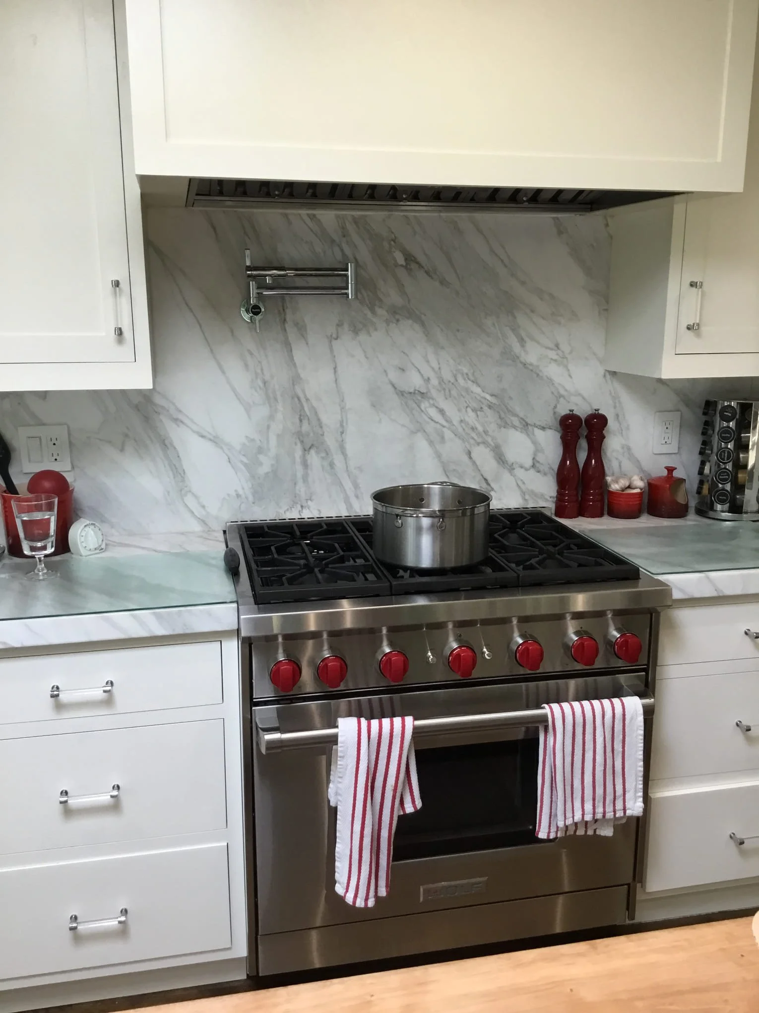 Stainless steel oven with red knobs in a white kitchen. Red and white striped towels hanging from the oven handle. White cabinets with silver knobs, marble backsplash, and kitchen counter. Red pepper grinders, garlic, and coffee capsules on the counter.
