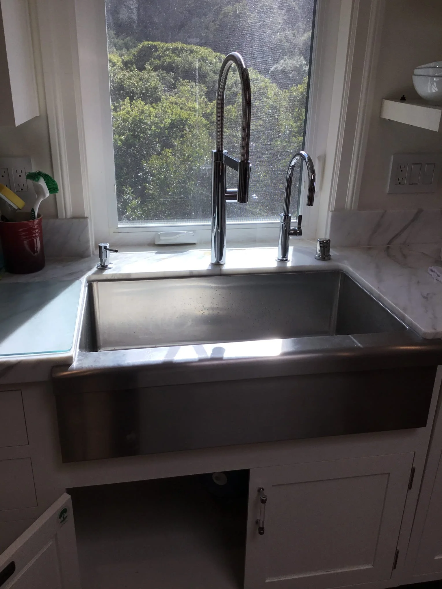 Kitchen sink area with a window showing trees outside. The sink has a modern pull-down kitchen faucet, a smaller secondary faucet, and a soap dispenser. To the left, there is a utensil holder with cleaning brushes and a yellow sponge. To the right, part of a white dish with a bowl on top is visible. The cabinetry below the sink is open, showing storage space.