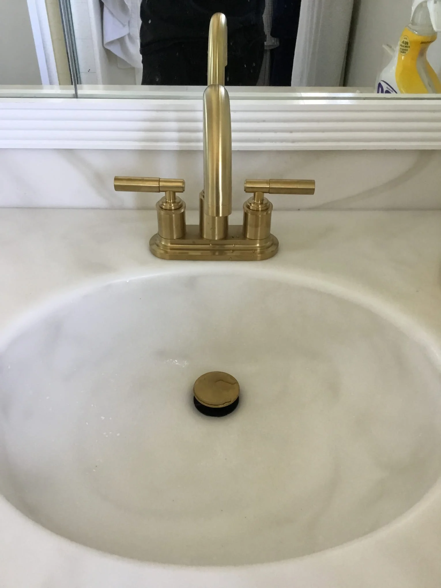 Bathroom sink with brass faucet, black and yellow drain stopper, and a bottle of cleaning spray visible in the mirror reflection.