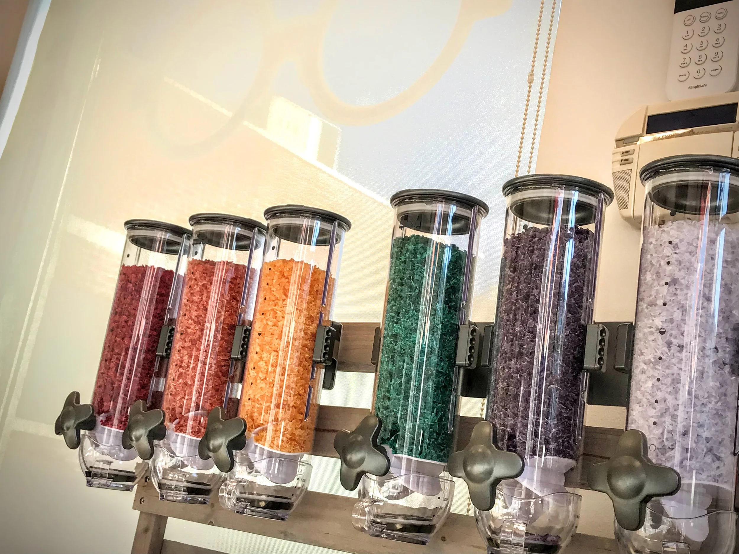 A row of six transparent dispensers containing acetate granules in different colors, attached to a metal frame.