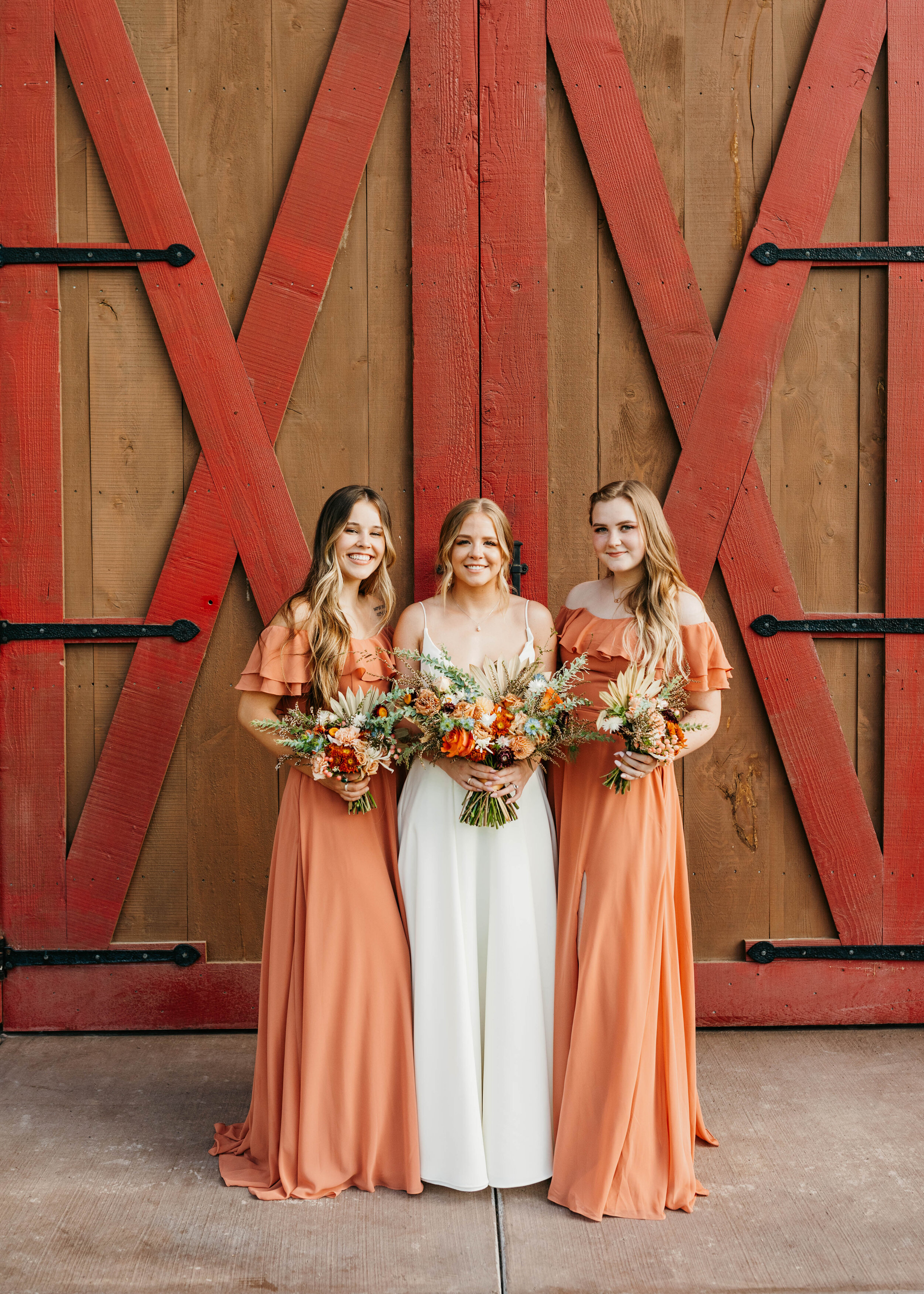  Posed in front of red and brown barn doors, this bride and her bridesmaids hold custom bridal bouquets by Cache Valley, Utah’s Sweet Afton Floral. orange peach bridesmaids dresses, orange off the shoulder bridesmaids dresses, custom wedding bouquets