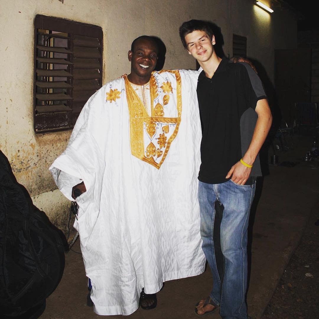 Toumani Diabaté and Josh Doughty standing together and smiling. Toumani Diabaté wears traditional West African dress, while Josh Doughty is dressed casually — a meeting of generations in the kora tradition.