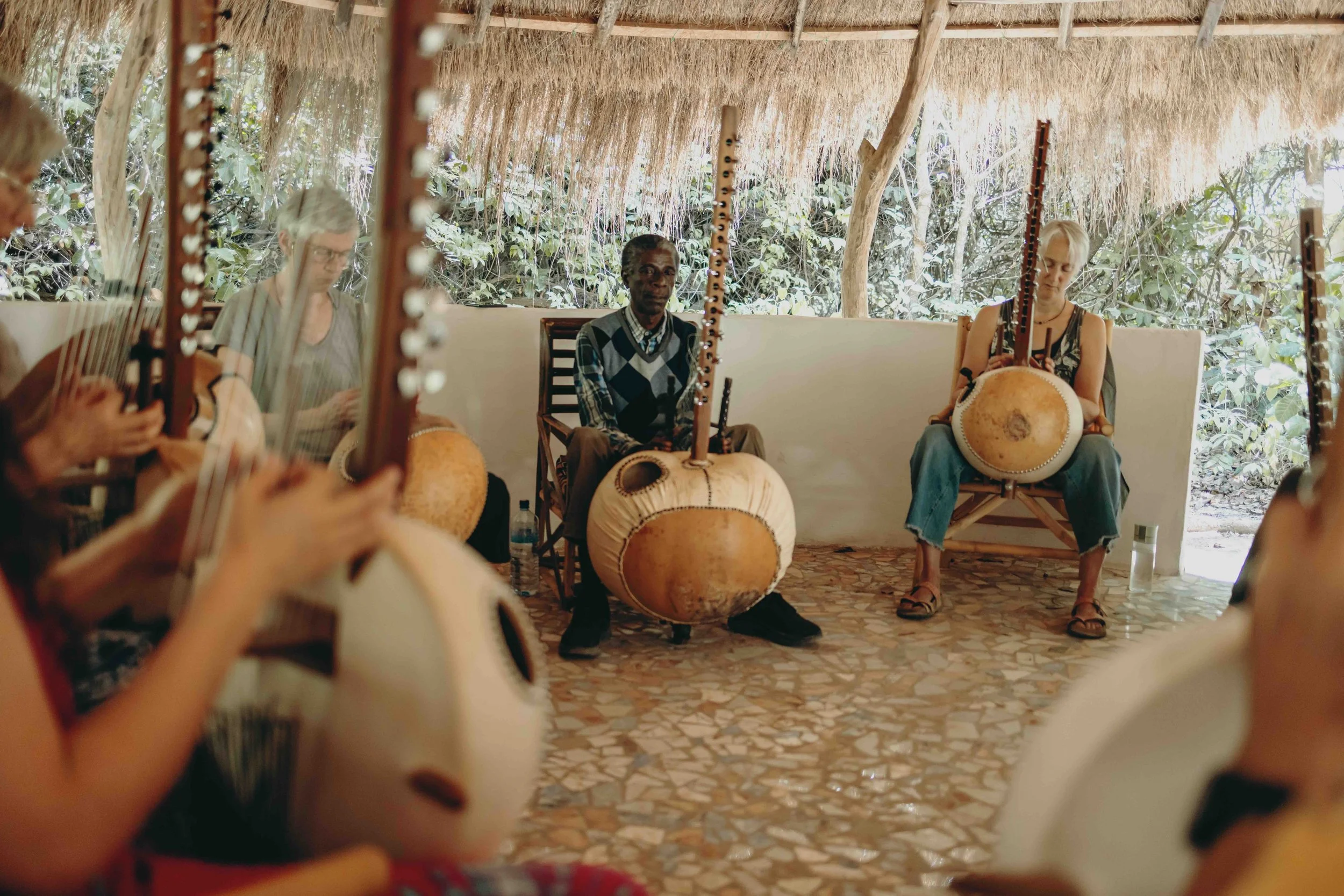 A guest teacher leading a kora session at one of Josh Doughty’s workshops, performing traditional West African music with a group of musicians in a thatched-roof setting surrounded by nature.
