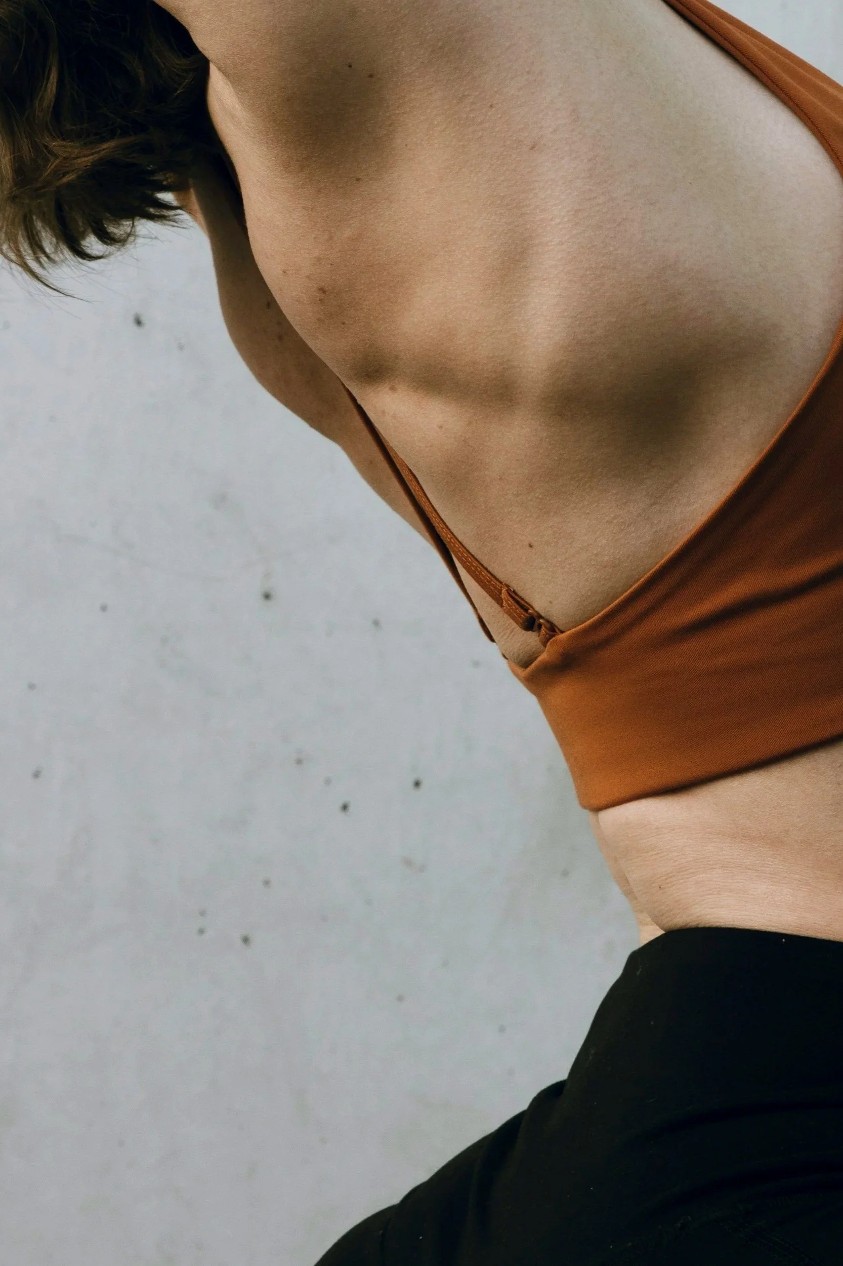 Image of the back of a woman wearing an orange yoga top and black yoga pants, standing in slight backbend position