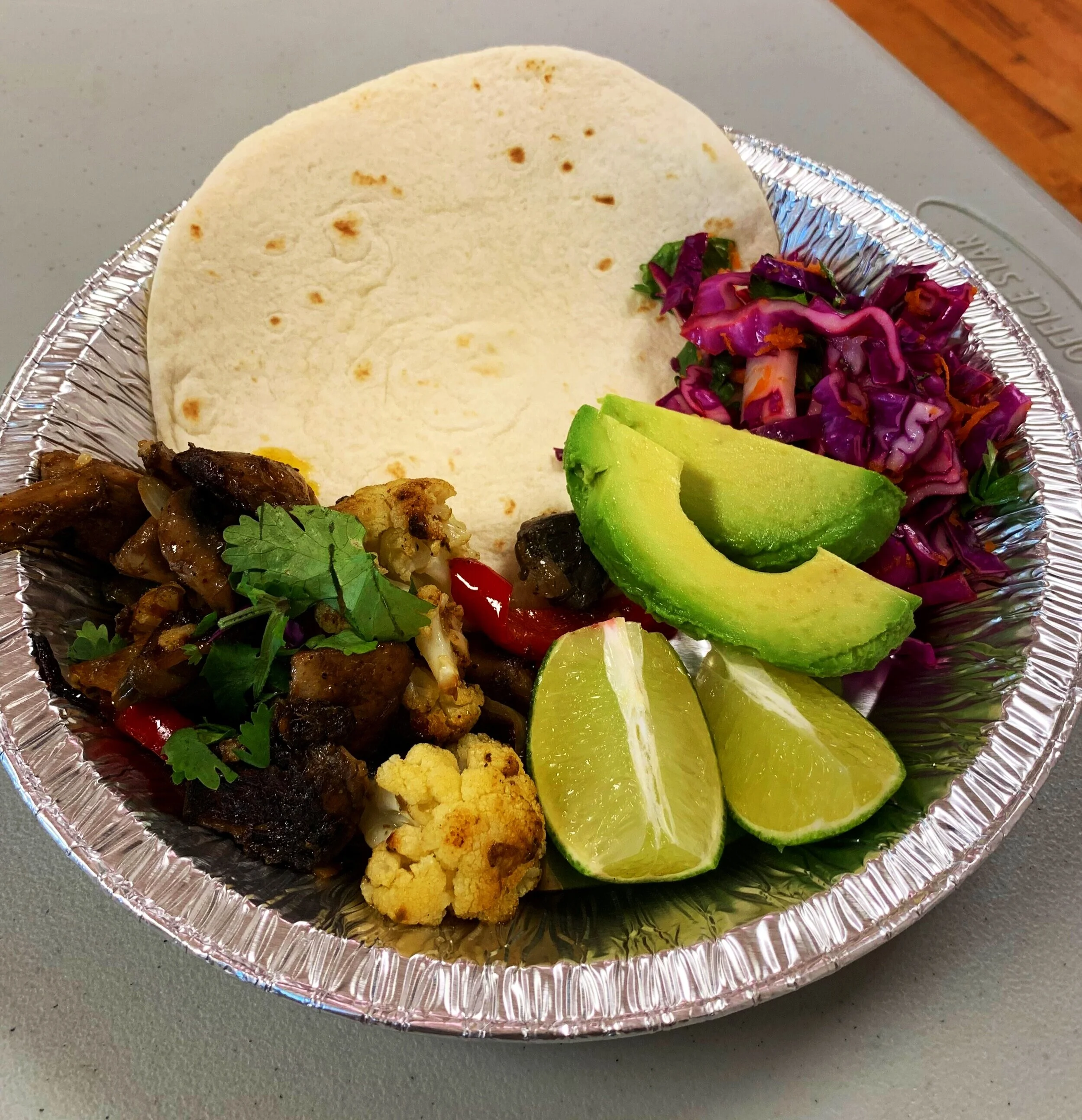 Picture of a silver foil plate containing limes, avocados, roasted cauliflower, red cabbage, a tortilla wrap, cilantro, red peppers and barbecued meat.