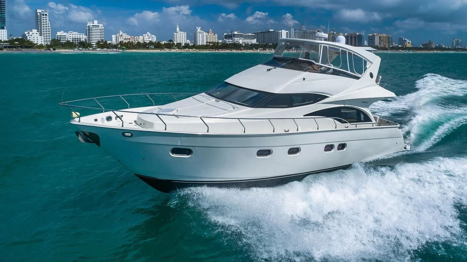 A white luxury motor yacht sailing on turquoise water with a city skyline in the background.