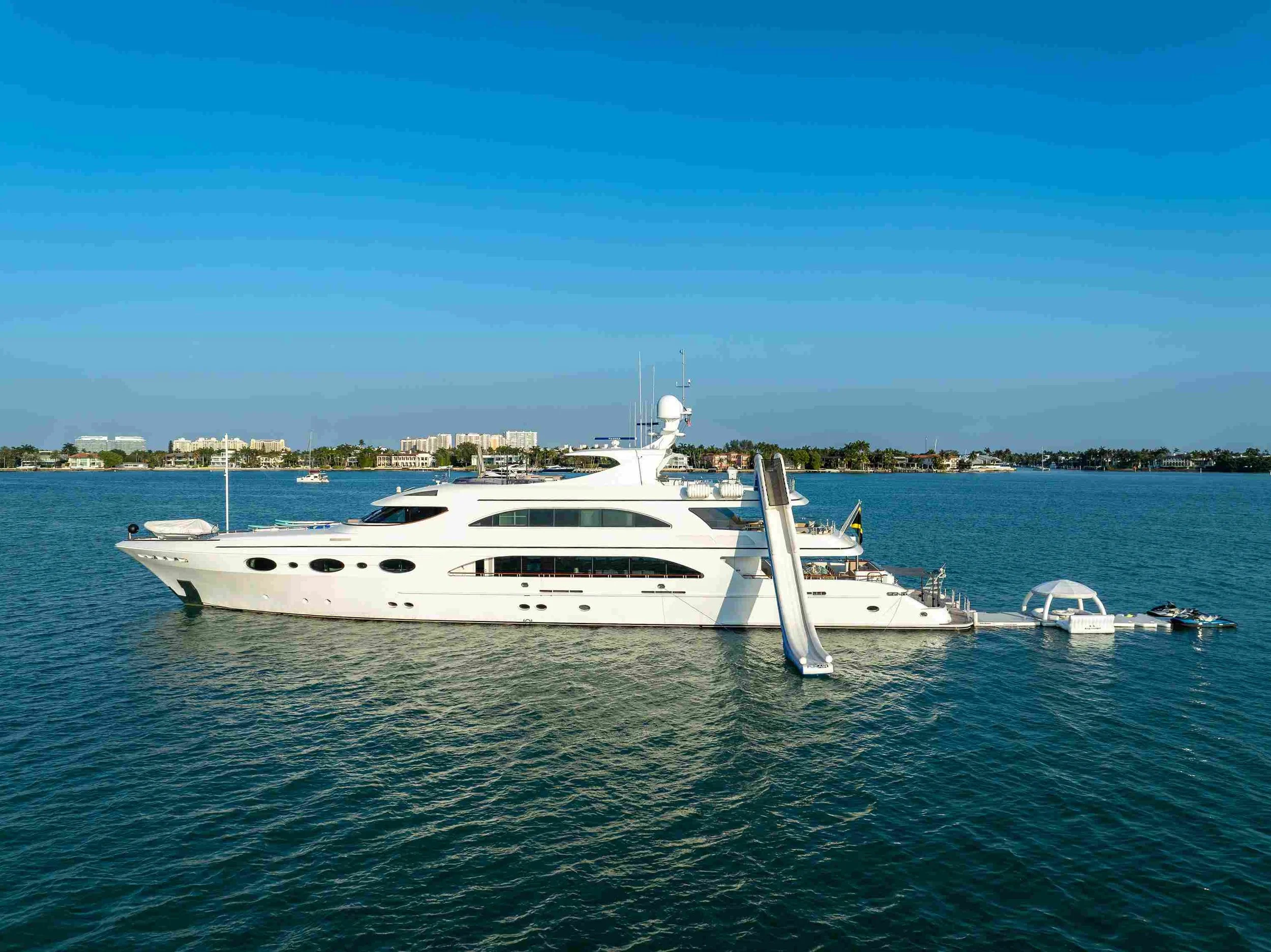 Luxury white yacht docked in a harbor with clear blue water and city buildings in the background on a sunny day.