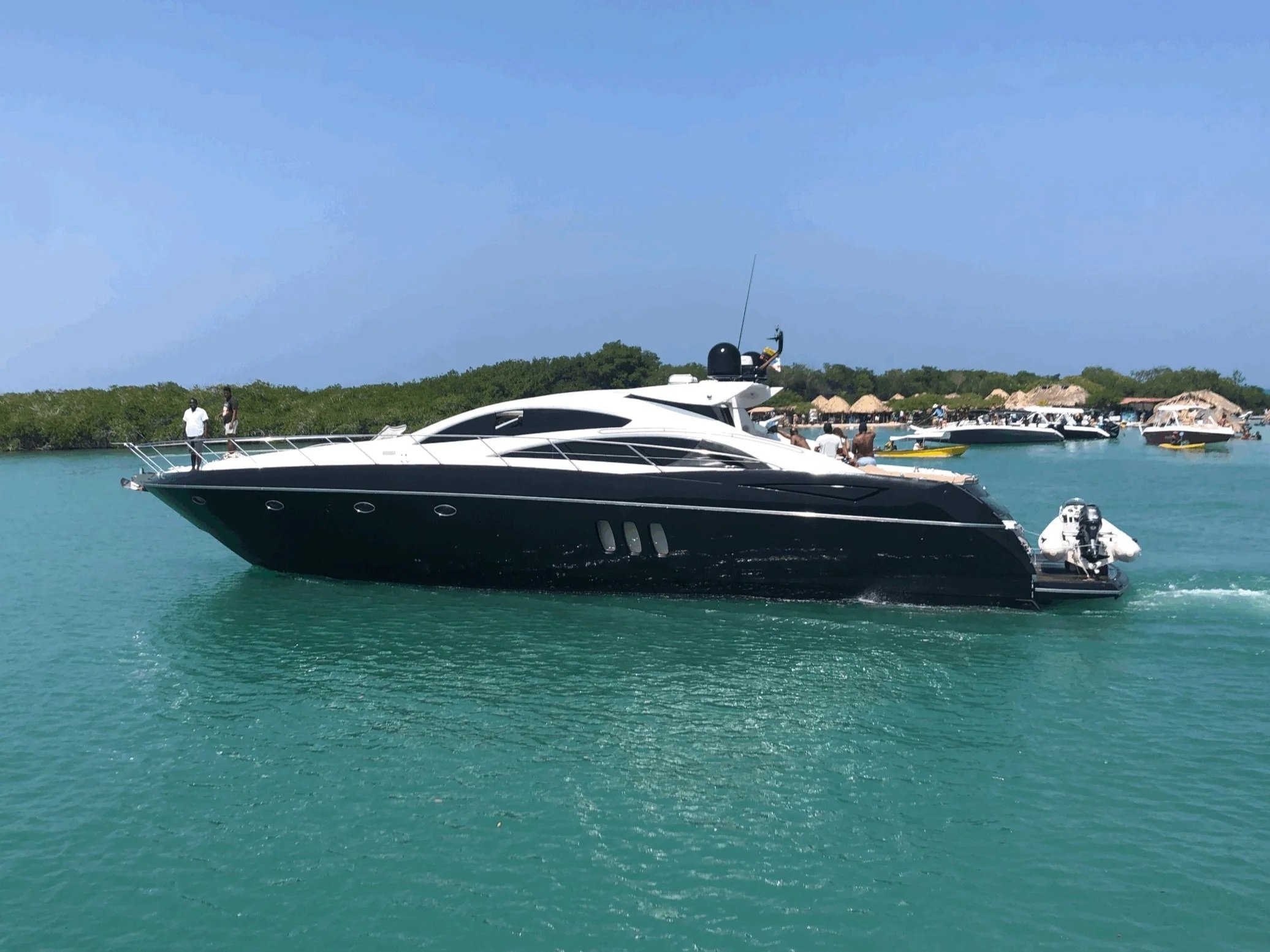 A large black and white yacht on calm turquoise water with a few people on board and around, and smaller boats in the background near a shoreline with lush greenery and thatched-roof structures.