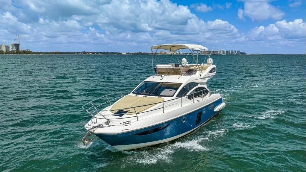 A large white and blue yacht cruising on the ocean with a city skyline and cloudy sky in the background.