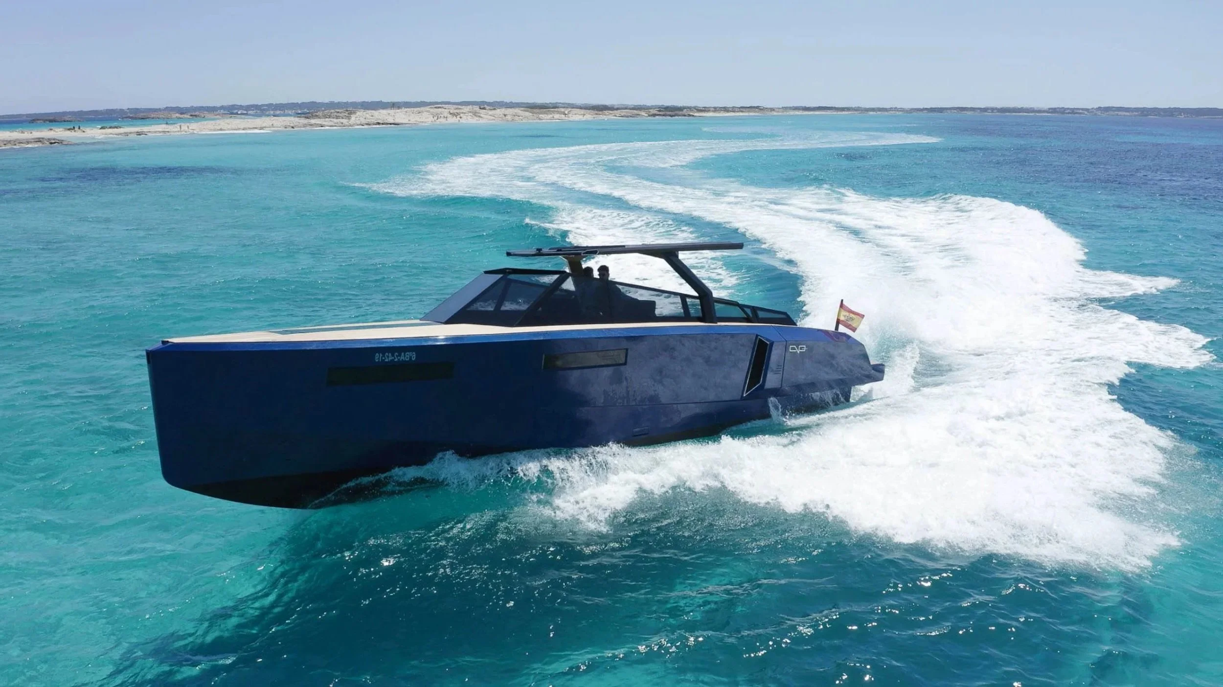 A sleek, dark blue boat speeding through turquoise ocean water, leaving a trail of white foam behind. The boat has a modern design with a glass cockpit and a small Spanish flag at the stern. In the background, a distant shoreline with rocky terrain and sparse vegetation is visible under a clear sky.