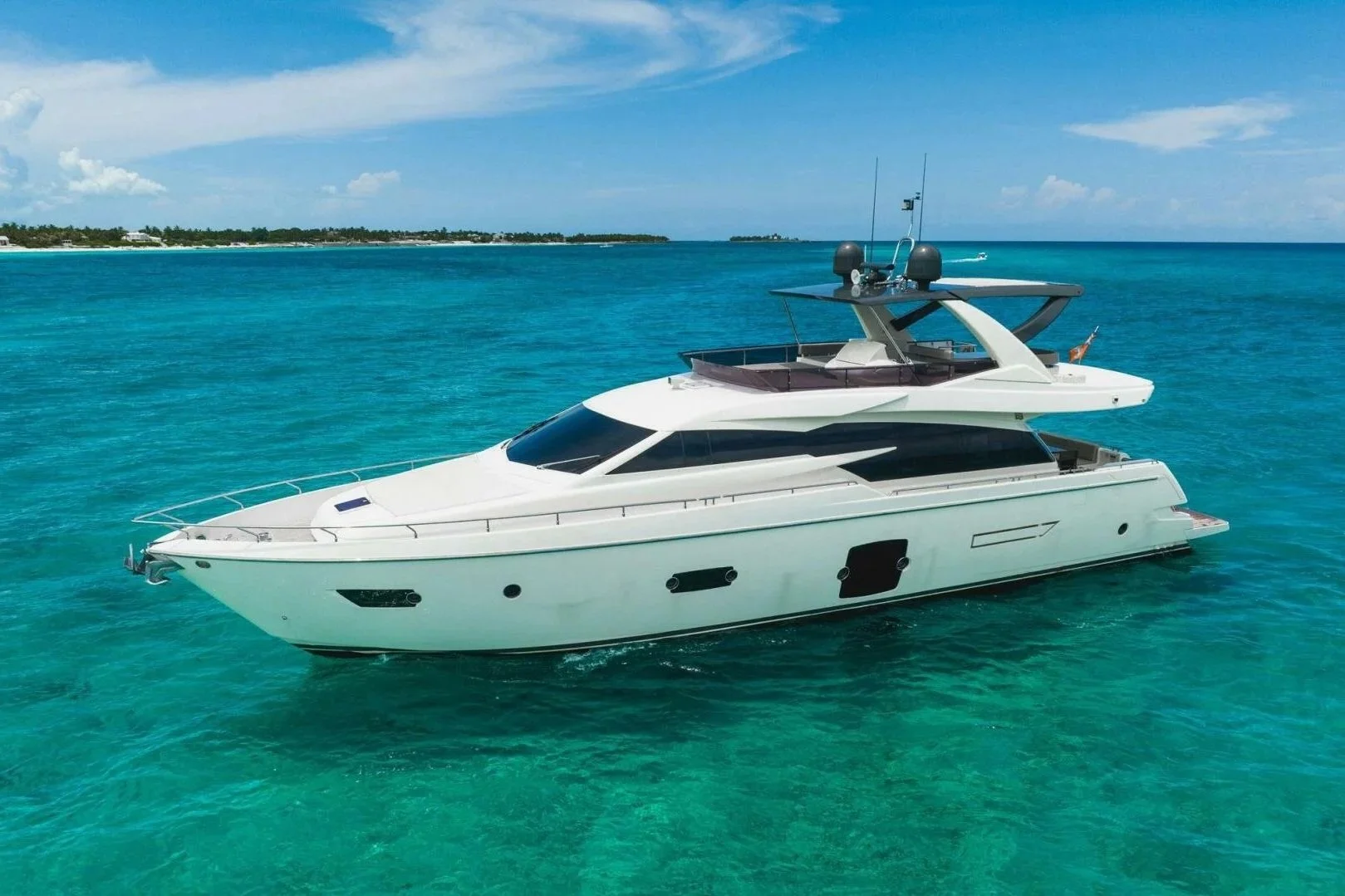 A white luxury yacht floating on clear turquoise ocean water with a distant shoreline and partly cloudy sky in the background.