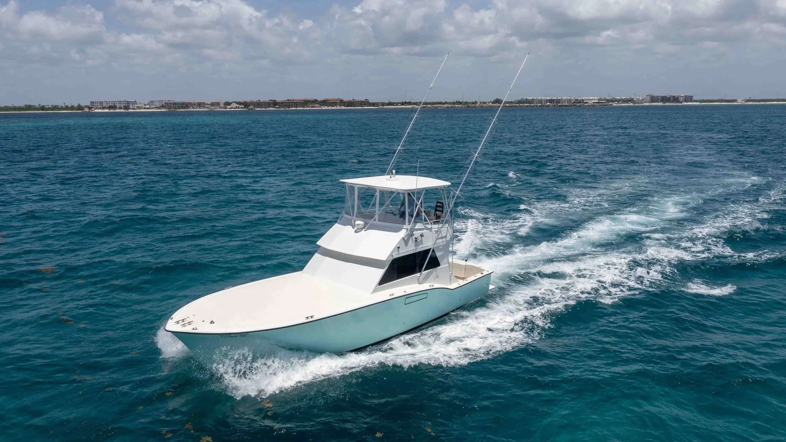A white motorboat cruises through blue ocean waters, leaving a wake behind. In the distance, there are buildings along a coastline under a partly cloudy sky.