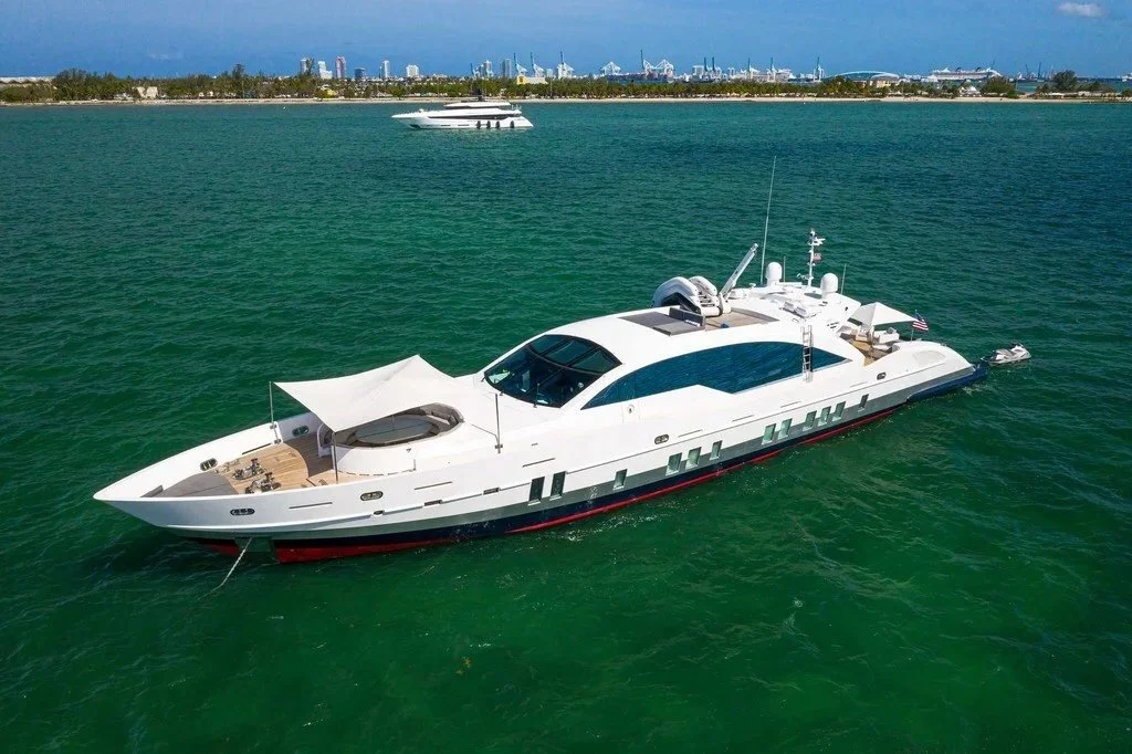 A large white yacht with a modern design floating on green water, with a shoreline and city skyline in the background.