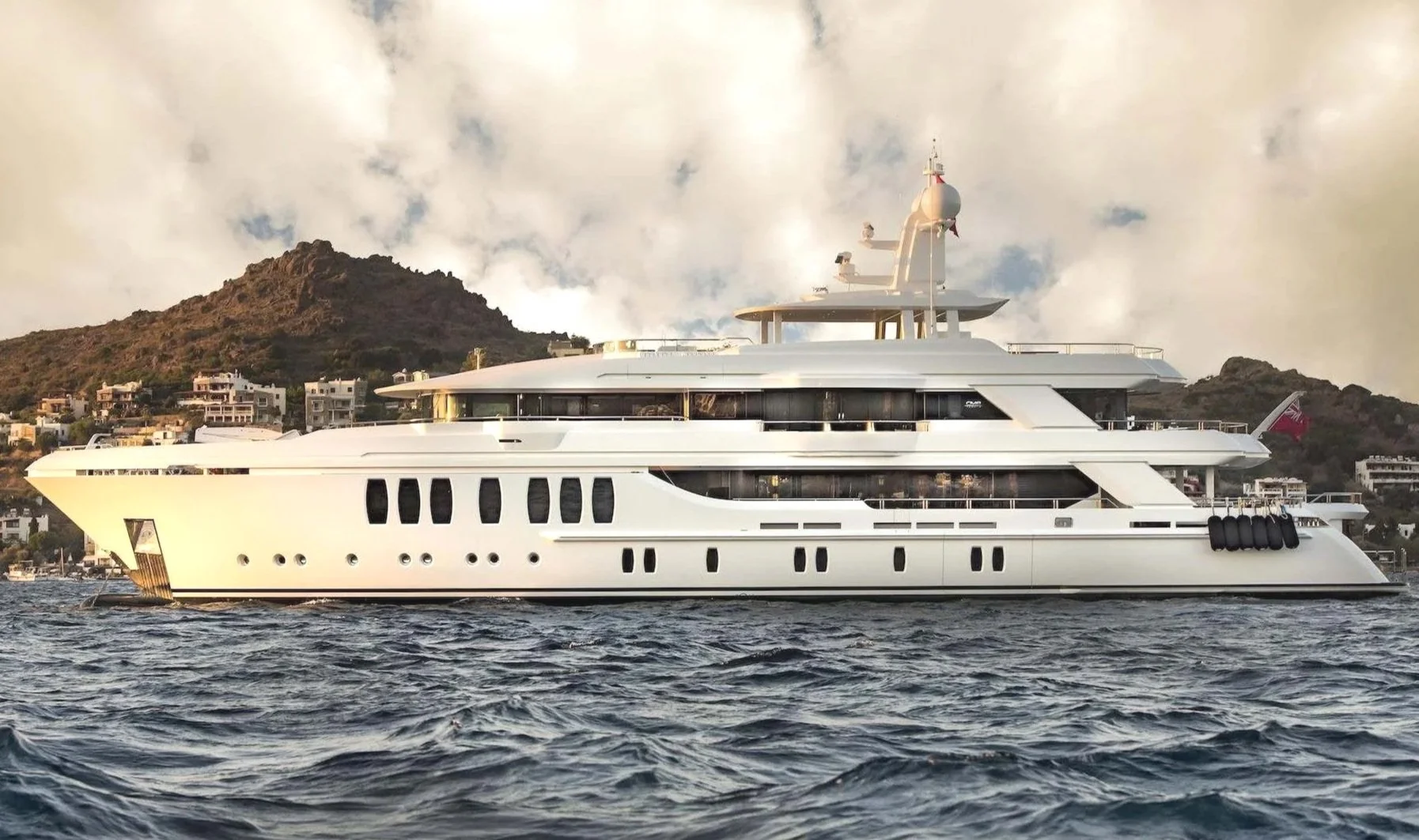 Large white luxury yacht on the water near a hilly coastline with residential buildings, under a cloudy sky.