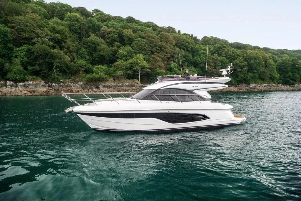 A white yacht with black accents floating near a rocky, forested shoreline.
