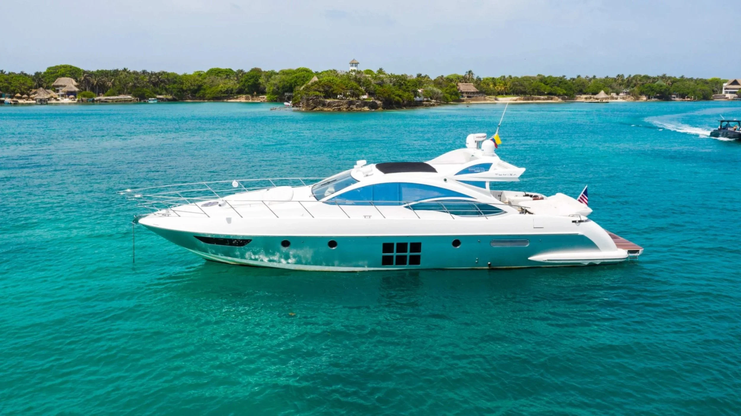 A luxury yacht floating on clear blue water near a tropical island with lush green trees and some beachfront structures.