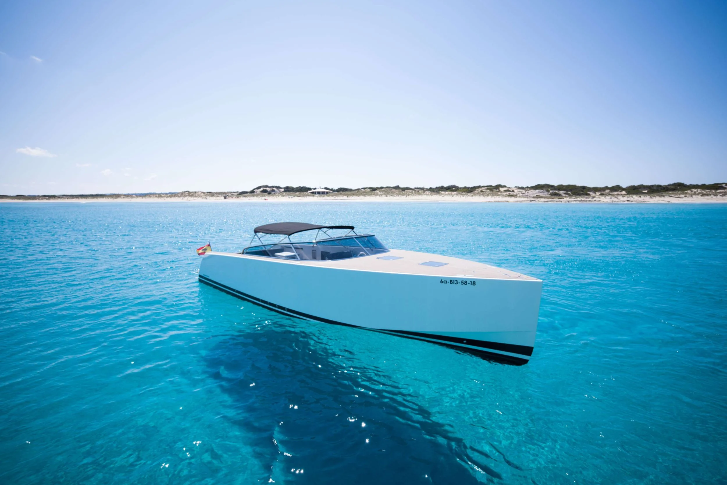 A white motorboat floating on clear blue water near a sandy shoreline under a bright, sunny sky.