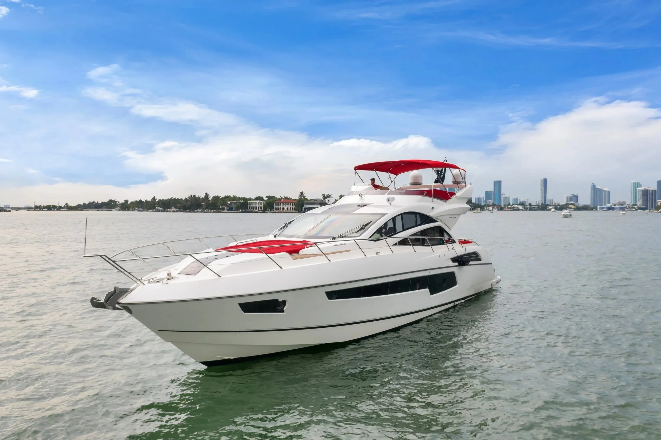 A luxury white yacht with a red canopy floating on calm water with a city skyline in the background under a partly cloudy sky.