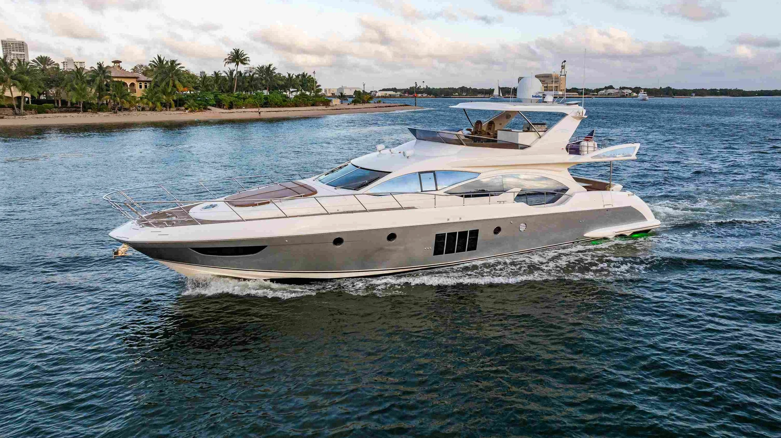 Luxury white yacht sailing on water near a shoreline with palm trees and buildings in the background.